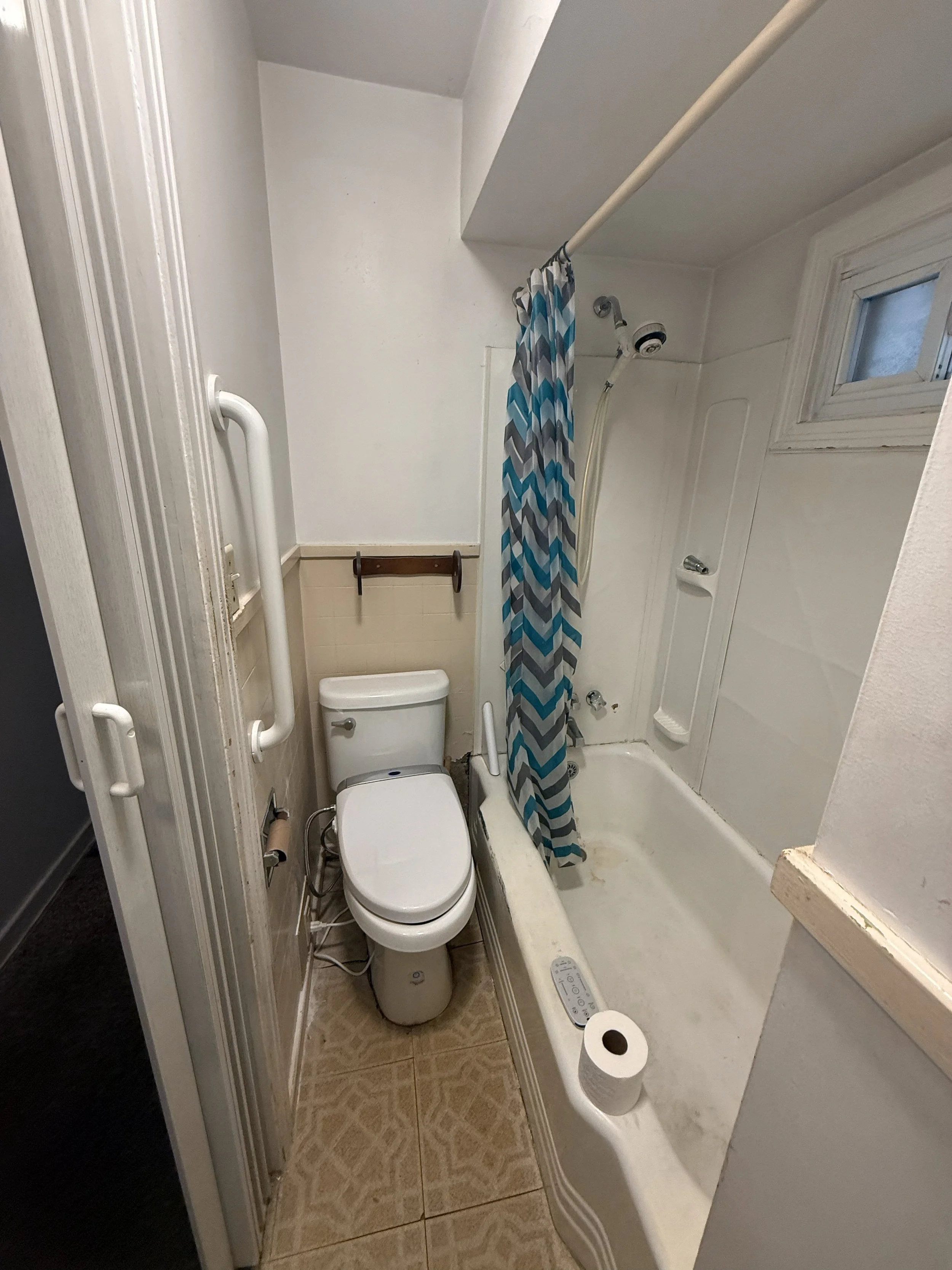 Before photo of a residential bathroom remodeling project in Vandergrift, Pennsylvania. This image shows the original bathroom featuring a fiberglass tub and shower, compact vanity, original toilet, vinyl flooring, and existing finishes prior to reno