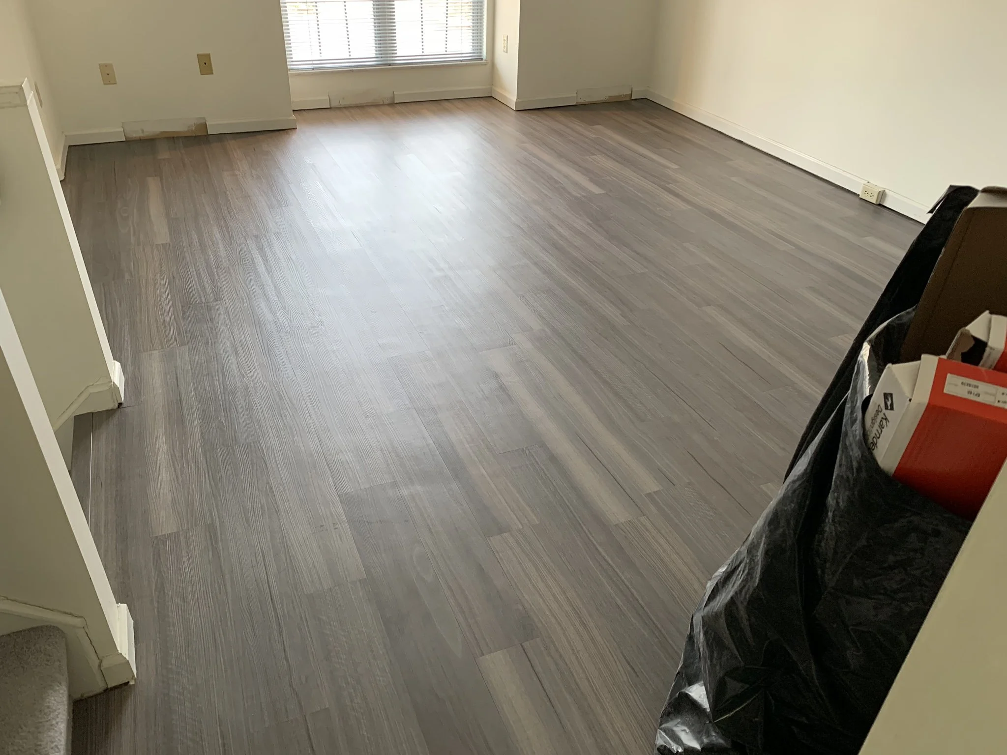 Empty room with gray wood flooring, beige walls, and a sliding glass door with blinds. There are some outlets and baseboards visible, and a black trash bag with a box inside toward the right of the image.