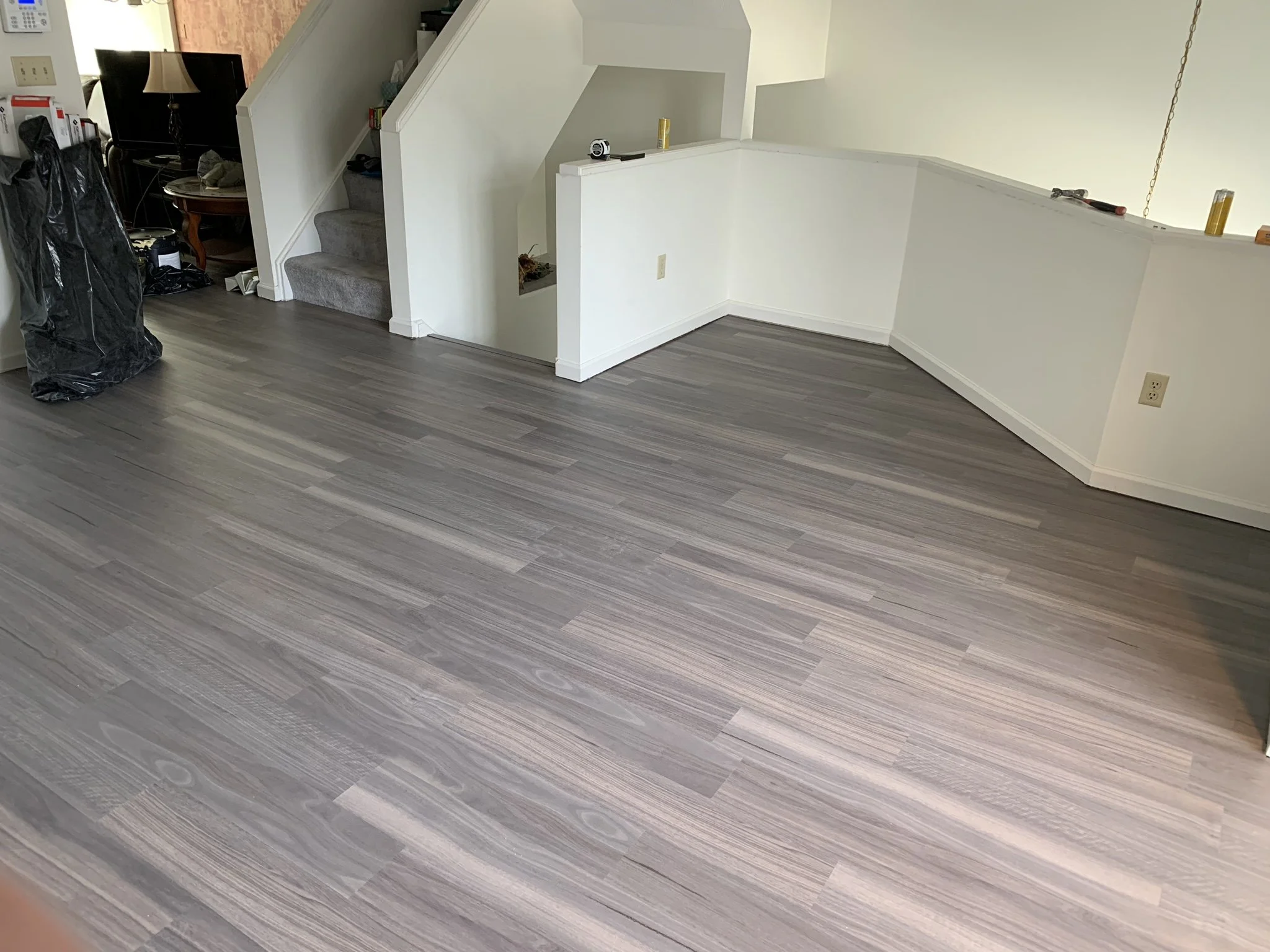 Living room with newly installed light gray wood flooring, stairs with carpet, small table, and various items near the wall.