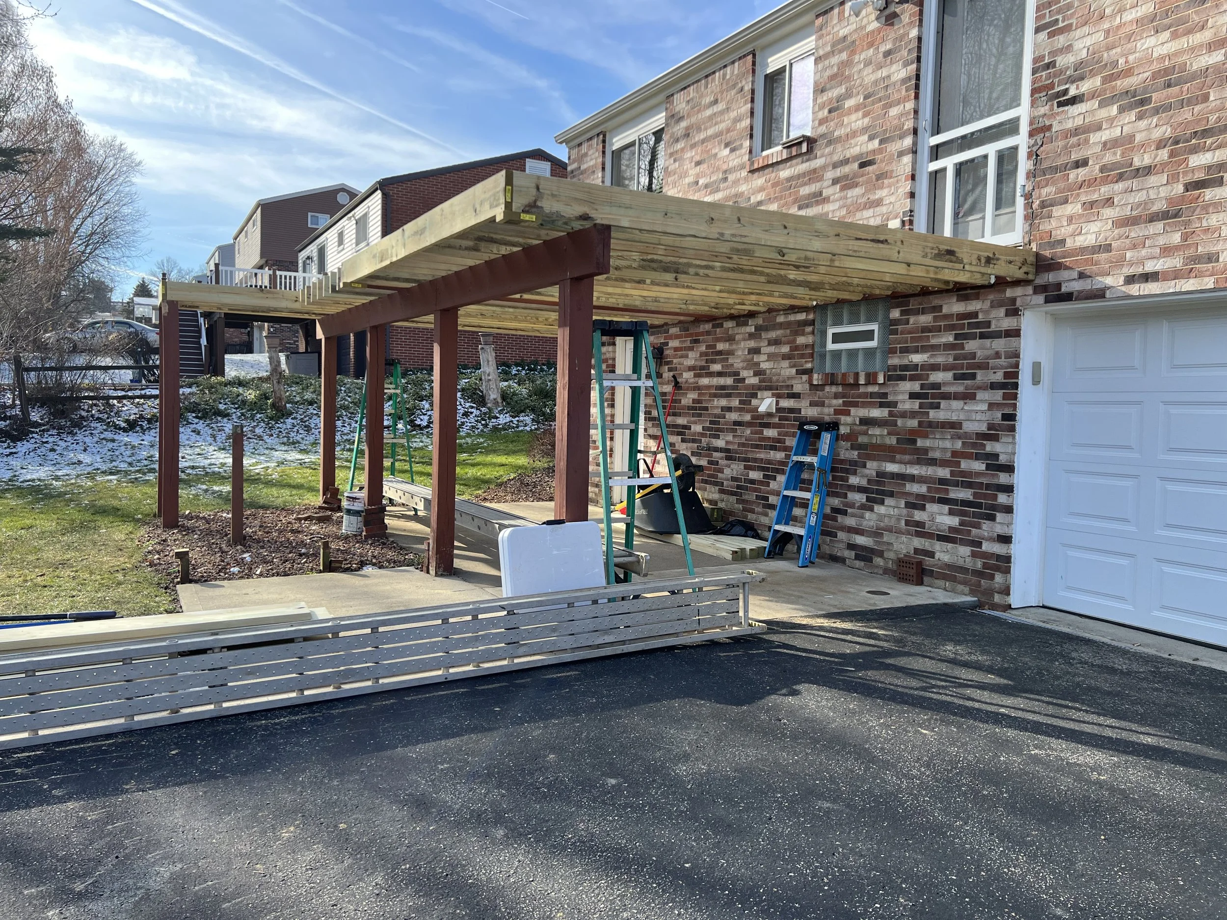Construction of a carport or patio cover next to a brick house with steps in the background. The structure has wooden beams and support posts, ladders and tools are present, and snow patches are on the ground.