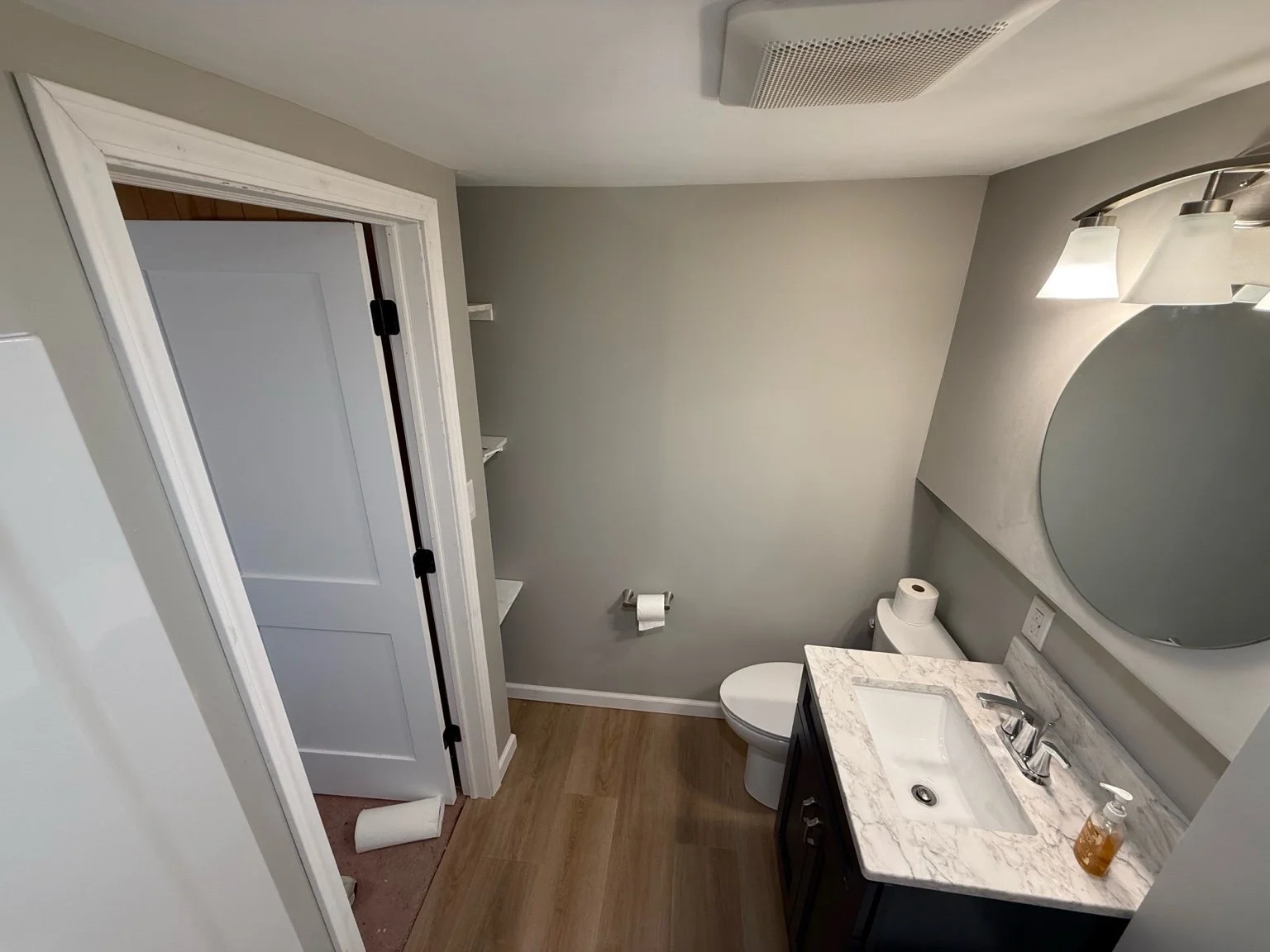 A small bathroom with a white toilet, a beige marble vanity with a small sink, a round mirror, a wall-mounted light fixture, and an open white cabinet with shelves. The bathroom has gray walls and wood-style flooring. There is a door slightly open, r