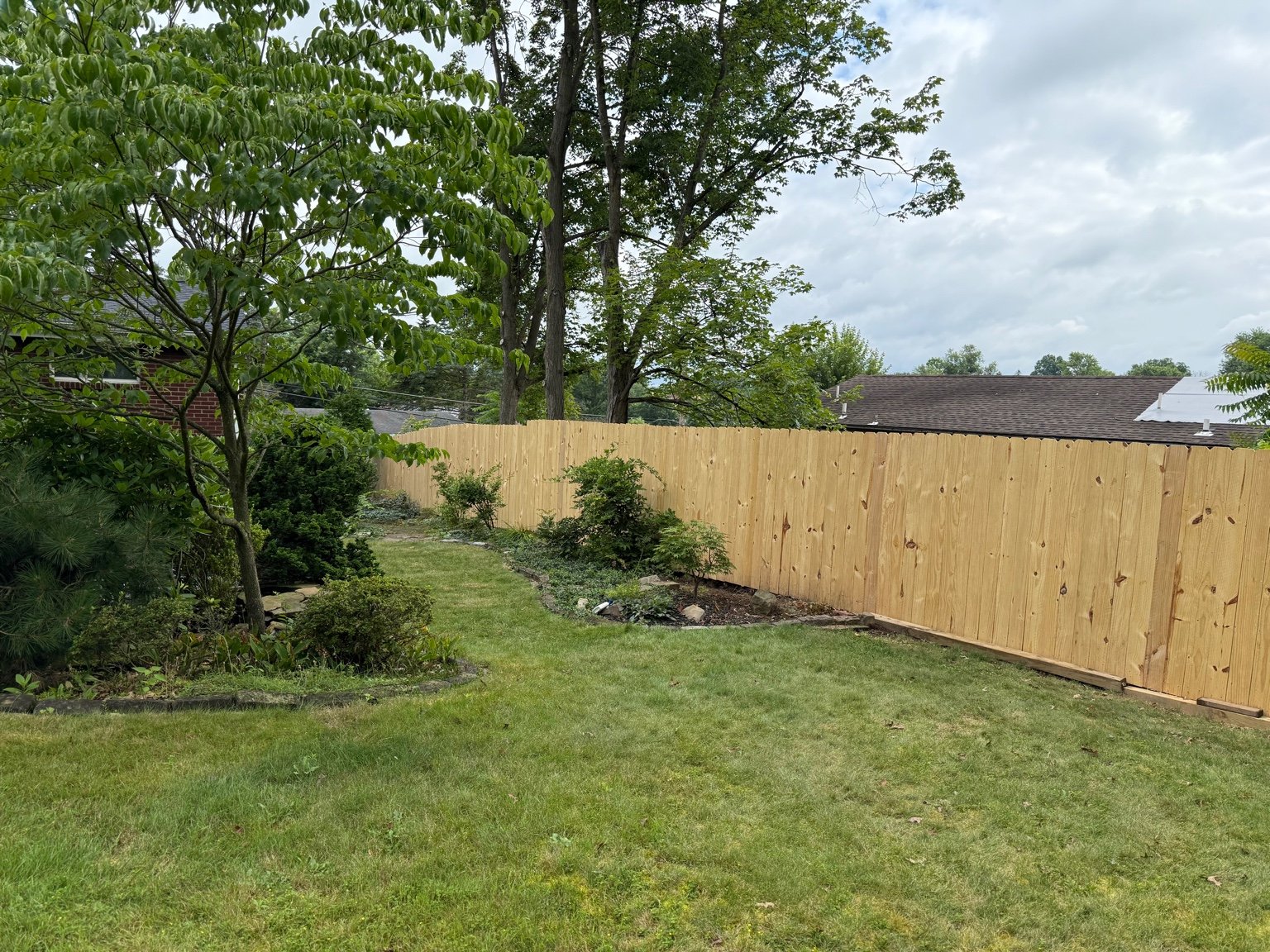 A backyard with a new wooden fence, green grass, trees, bushes, and some small plants along the edge of the fence.