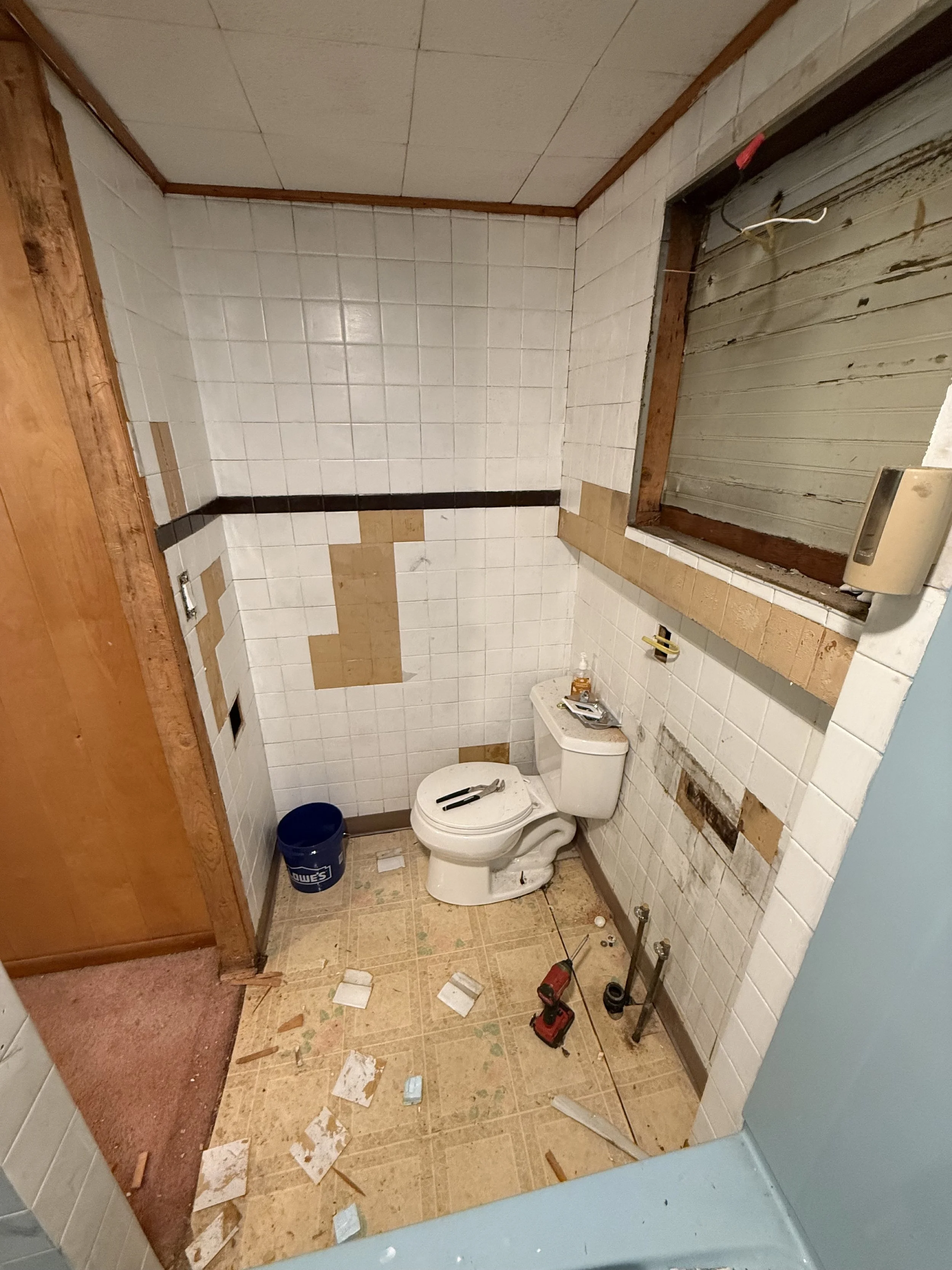 A small, partially renovated bathroom with a toilet, construction tools, and debris on the floor. The walls are tiled with white tiles, some of which are missing or damaged, and a window opening is visible without glass or window frame.