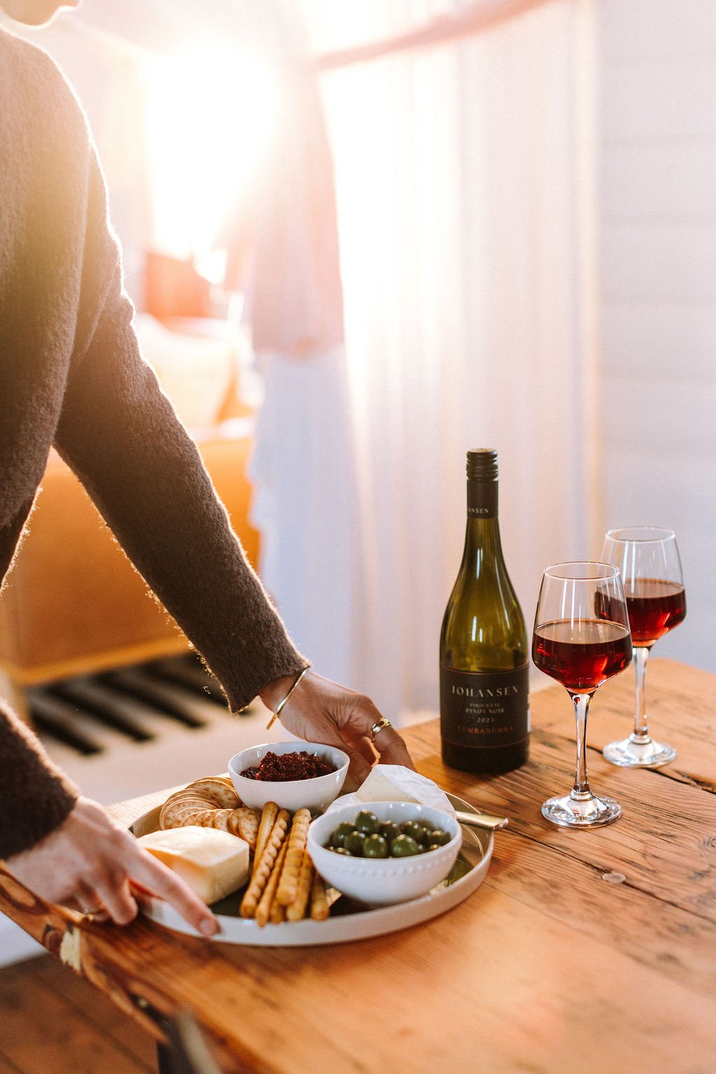 A person setting a wooden table with a tray of cheeses, crackers, green olives, and a bowl of what appears to be tapenade or fruit spread, with a bottle of red wine and two glasses of red wine nearby, in a warmly lit room.