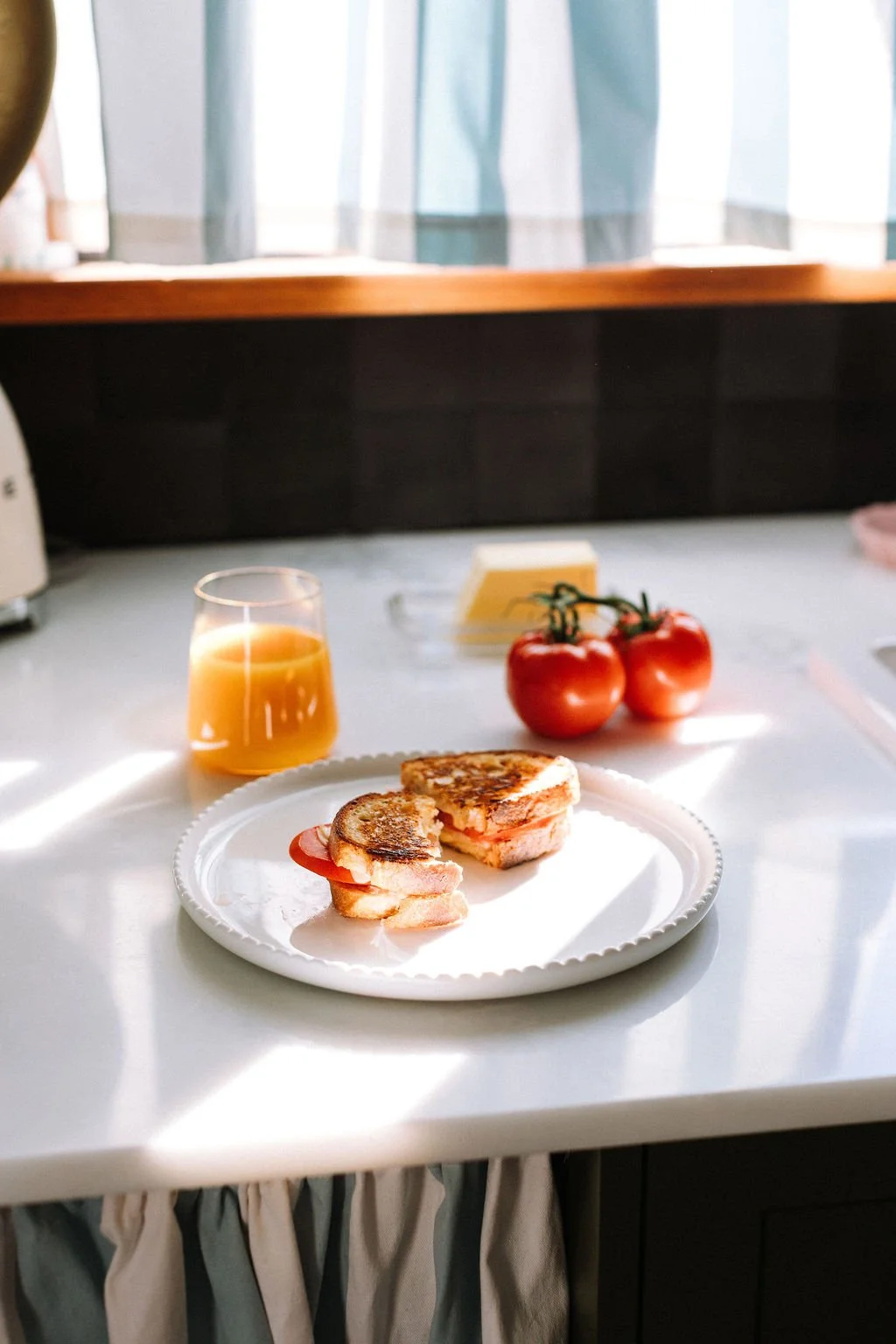 A plate with a grilled sandwich, a glass of orange juice, tomatoes, and a small stack of butter on a white kitchen counter.
