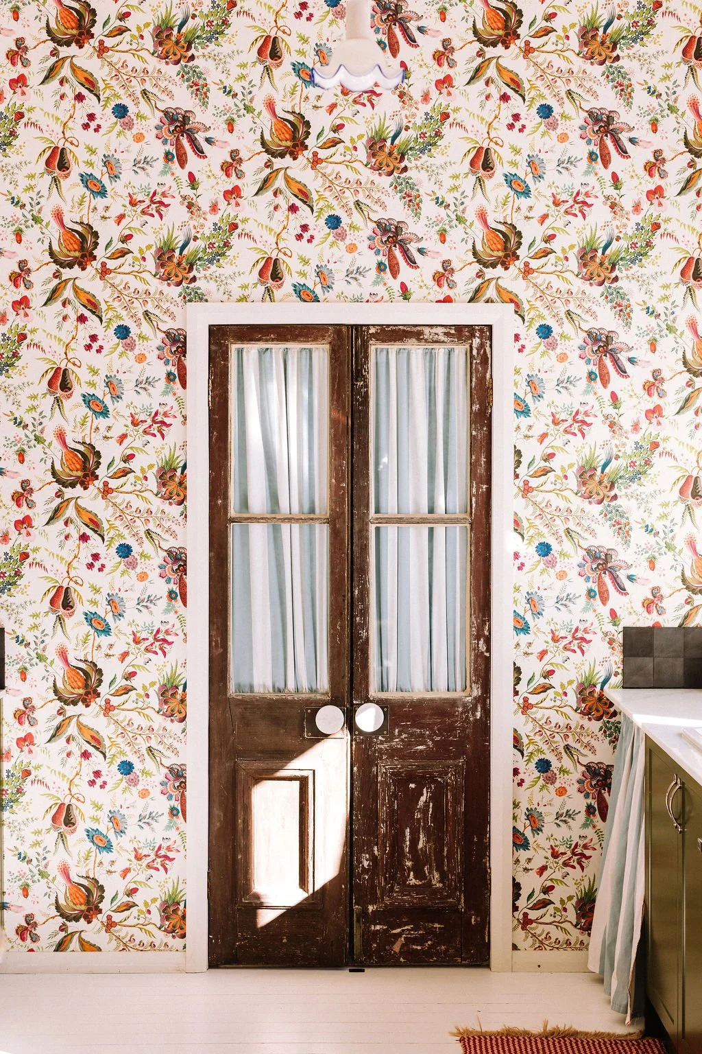 A pair of vintage wooden doors with glass panes and white curtains, set in a doorway with colorful floral wallpaper in the background.