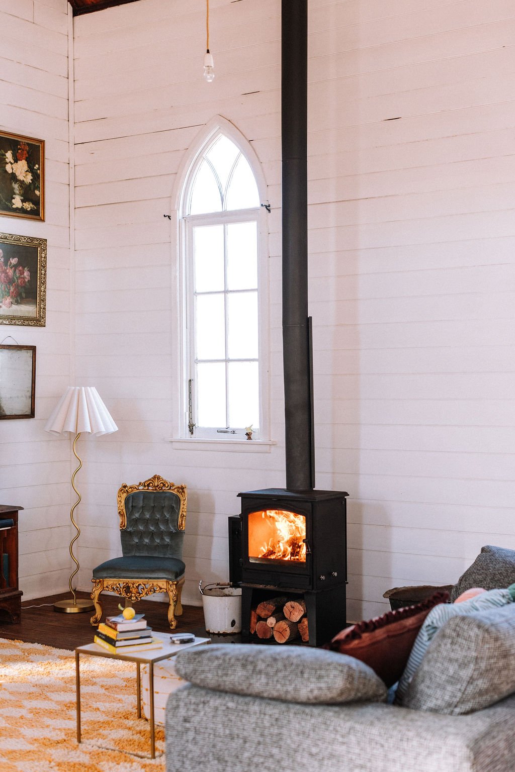 Cozy living room with a wood stove burning, grey couch, ornate armchair, small coffee table with books, a white lamp, and a window with sunlight, in a white-paneled wall space.