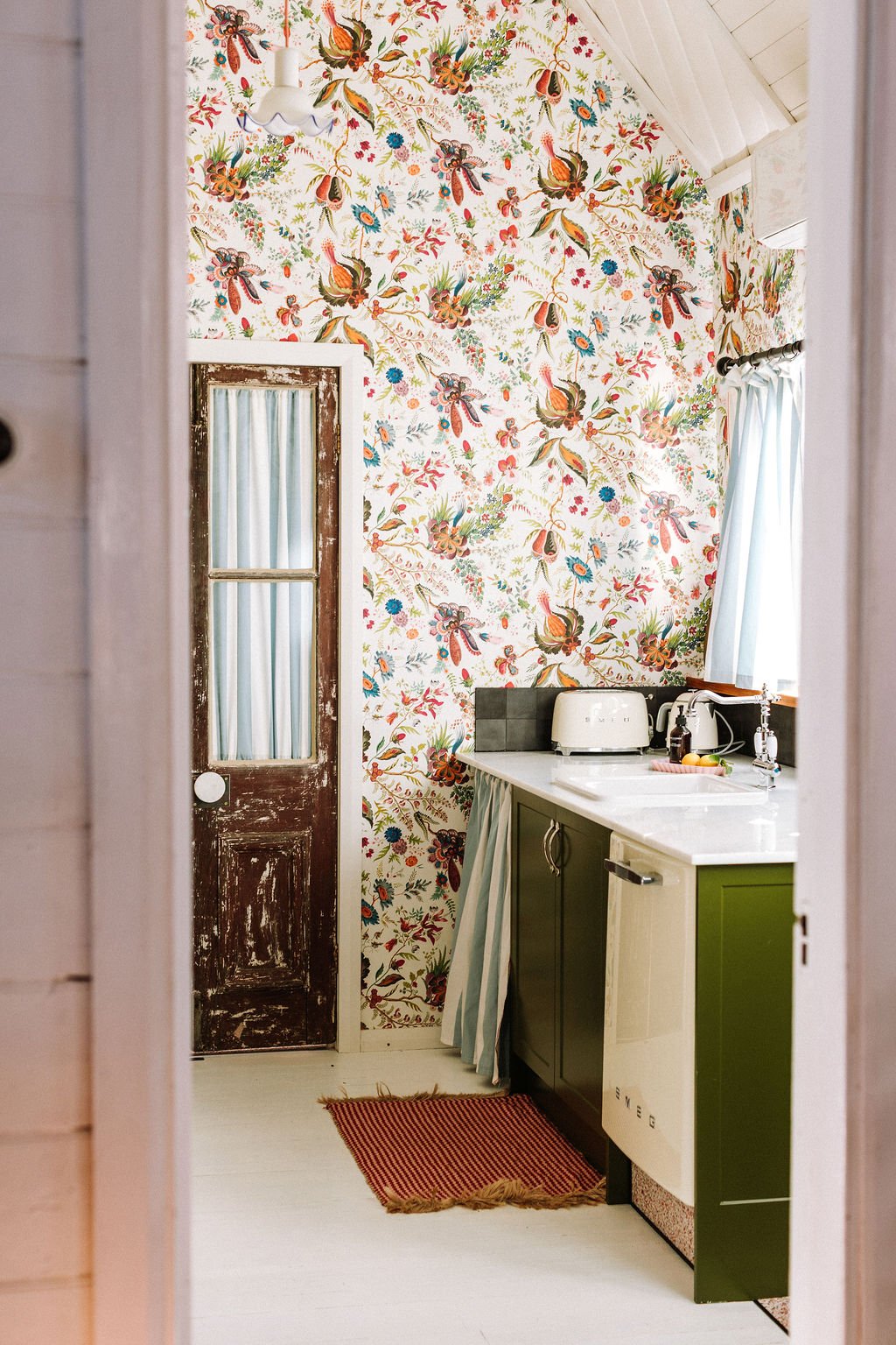 A cozy kitchen with wallpaper featuring a colorful floral and bird pattern, a white ceiling, and a window with light blue curtains. There is a rustic wooden door, a small white countertop with a toaster, kettle, and soap dispenser, a green cabinet, a