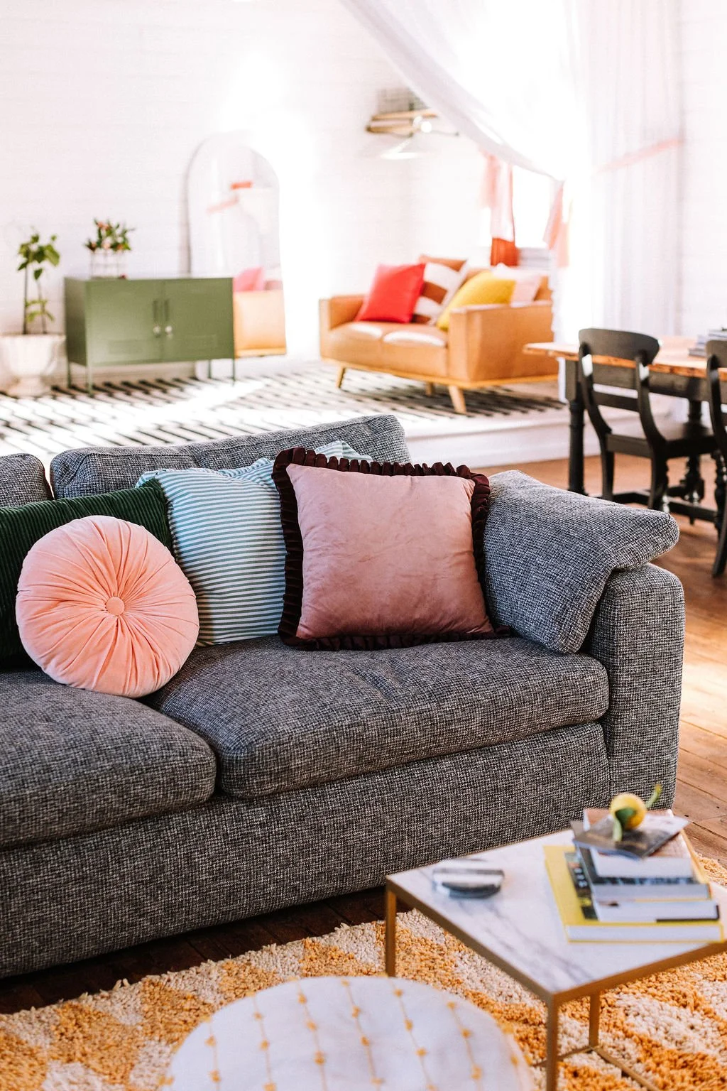 A cozy living room with a gray upholstered sofa decorated with colorful pillows, including pink, green, and striped ones. There is a small white marble coffee table with books and a lemon in the foreground. In the background, a bright room features a