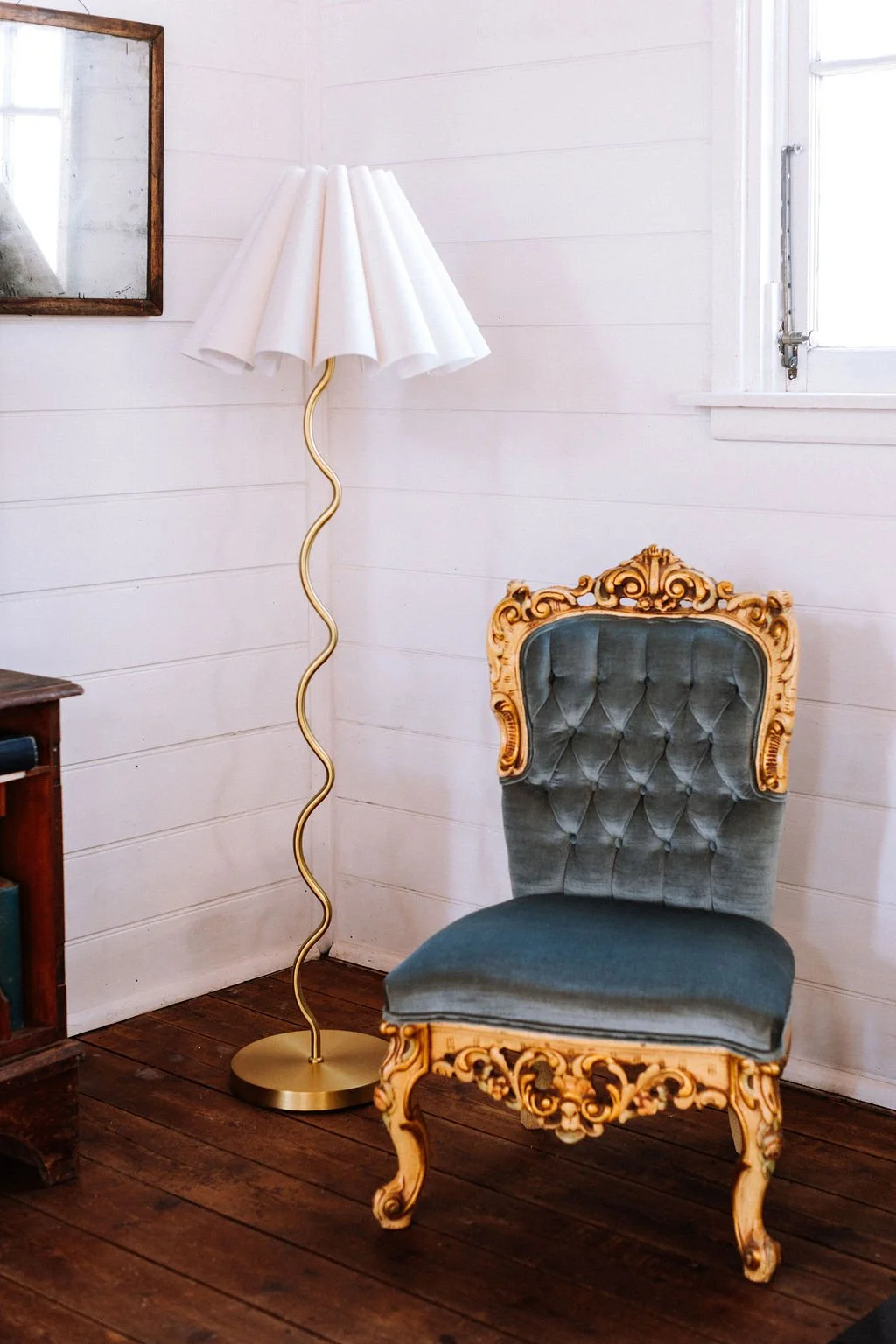 A vintage-style grey upholstered armchair with ornate wooden carvings, and a modern floor lamp with a wavy gold stand and a white fabric shade, set against white wooden wall paneling and a wooden floor.