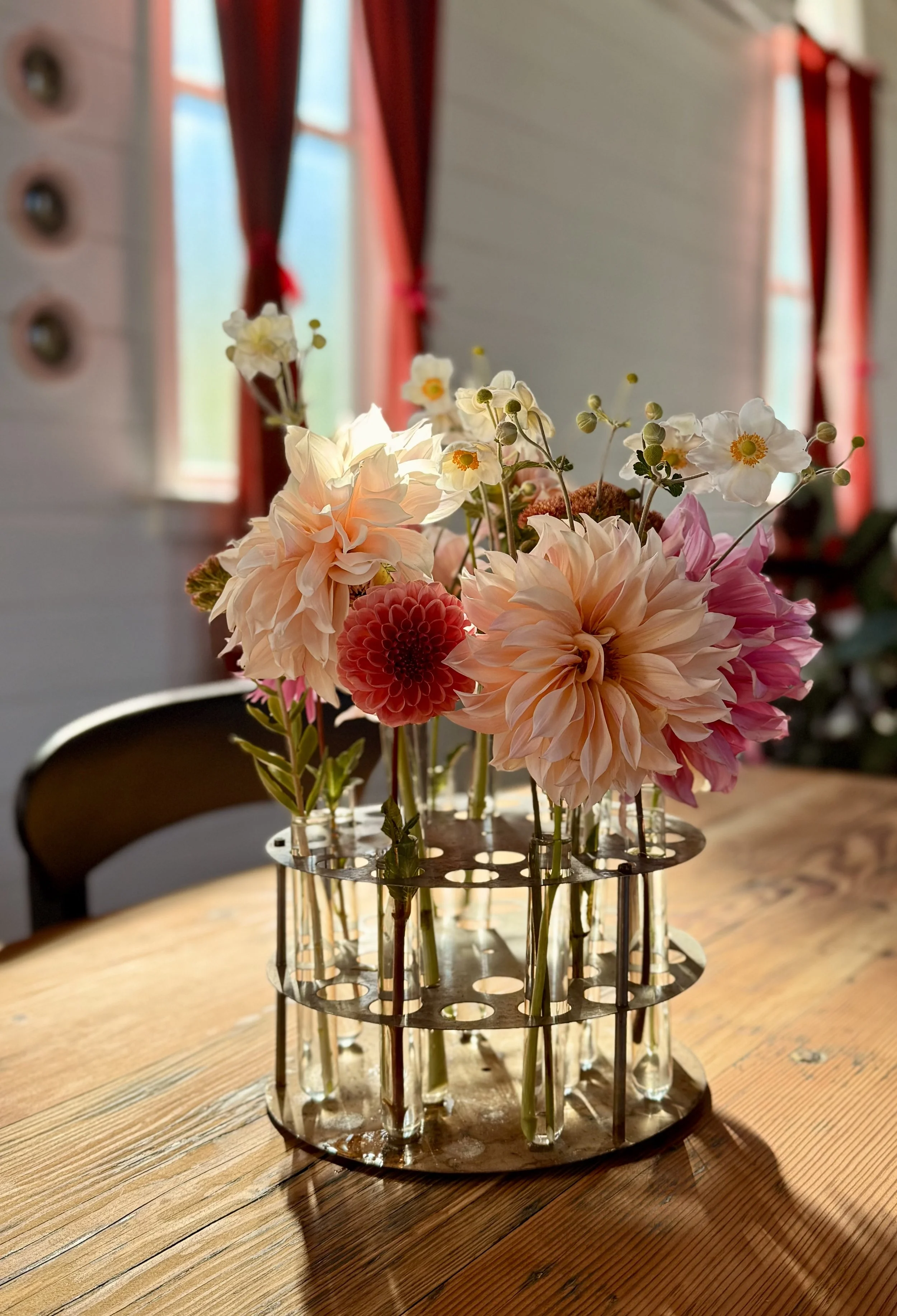 Flowers in a metal flower holder on a wooden table, with sunlight streaming through a window.