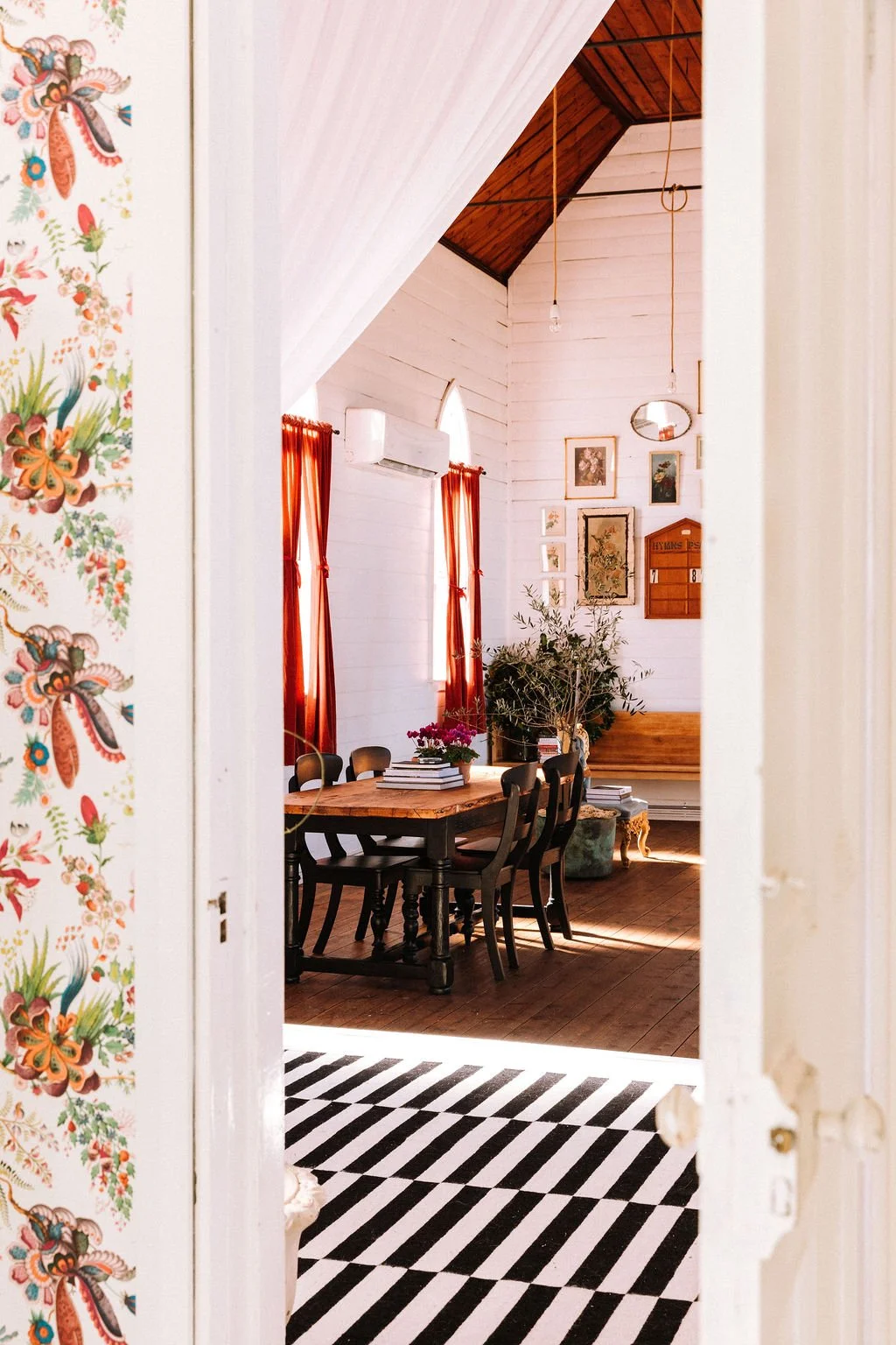 View through doorway into a cozy dining area with wooden table and black chairs, adorned with a pink flower arrangement, warm red curtains, framed artwork on white wooden walls, and plants, with sunlight coming in.