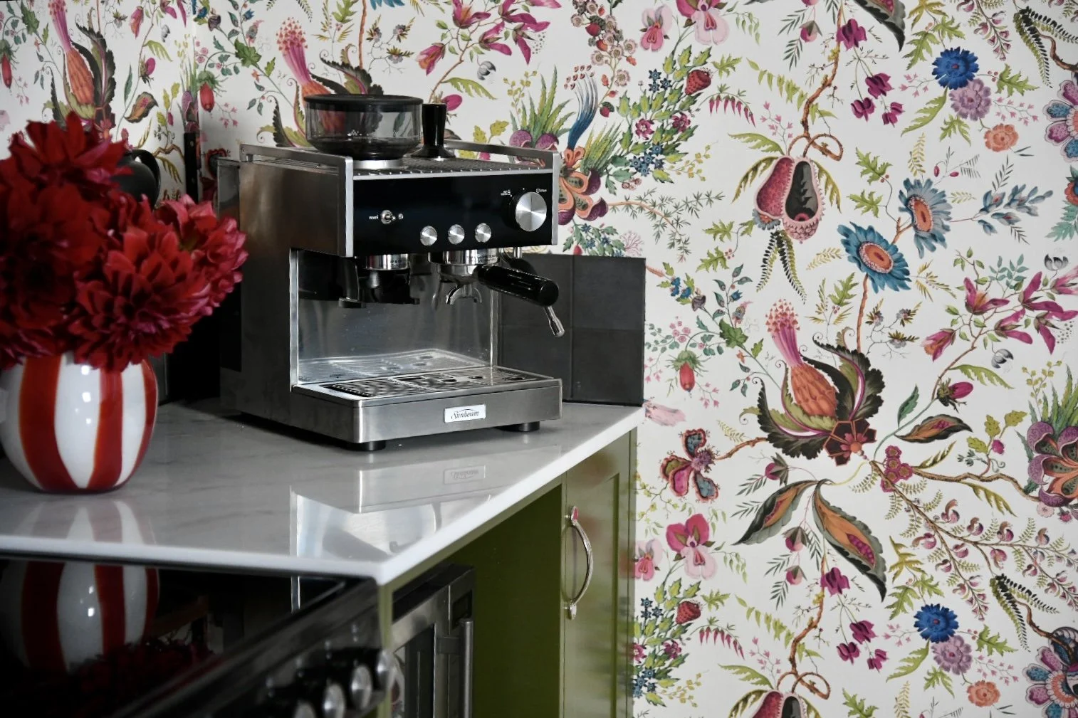 Espresso machine on marble countertop with colorful floral wallpaper and red flowers in a white and red striped vase.