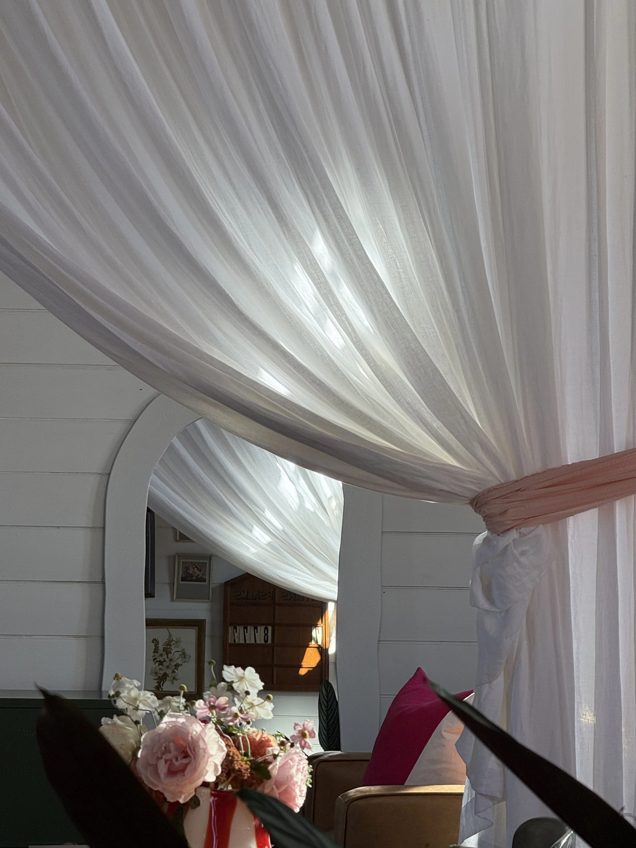 Room with white draped curtains, a mirror reflecting the scene, a pink pillow on a chair, and a bouquet of pink and white flowers.
