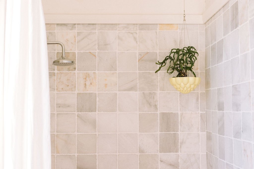 A shower with a hanging potted plant in a yellow holder in a bathroom with beige tiled walls.
