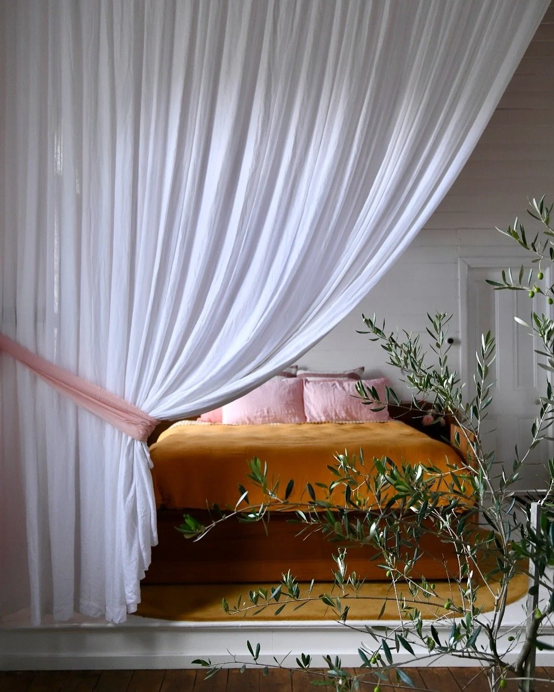 Cozy bedroom with a canopy bed, white drapes, yellow bedding, pink pillows, and a potted plant.