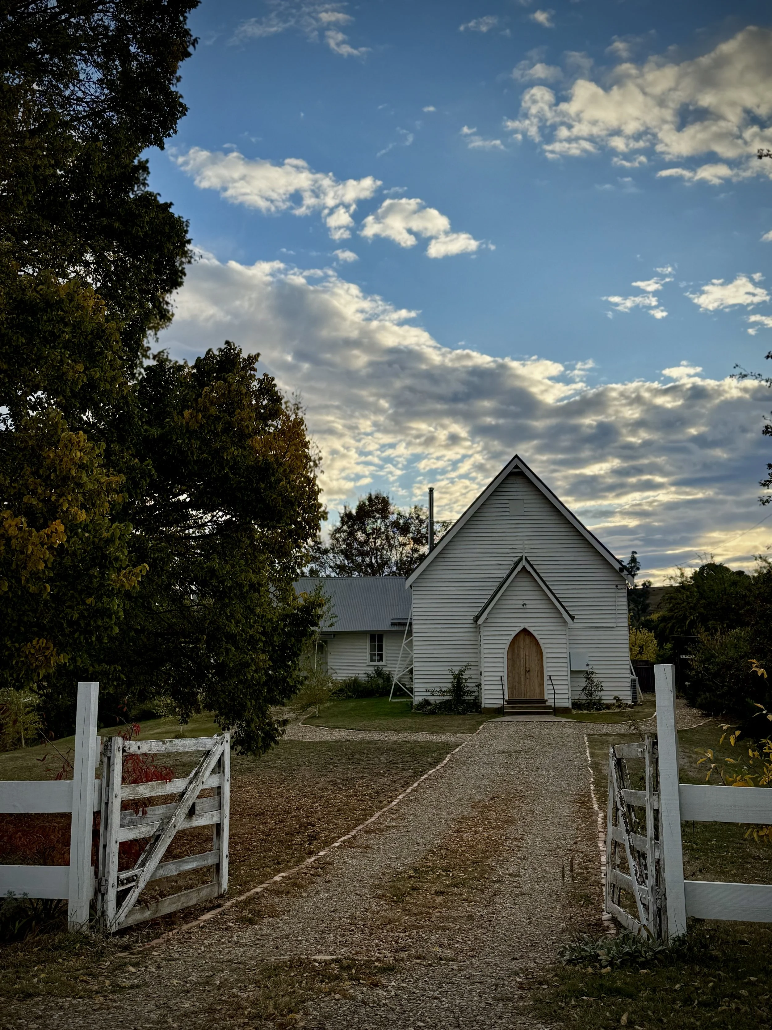 A rustic white chapel with a pointed roof sits at the end of a gravel path, flanked by an open wooden gate and surrounded by trees, under a sky filled with scattered clouds.