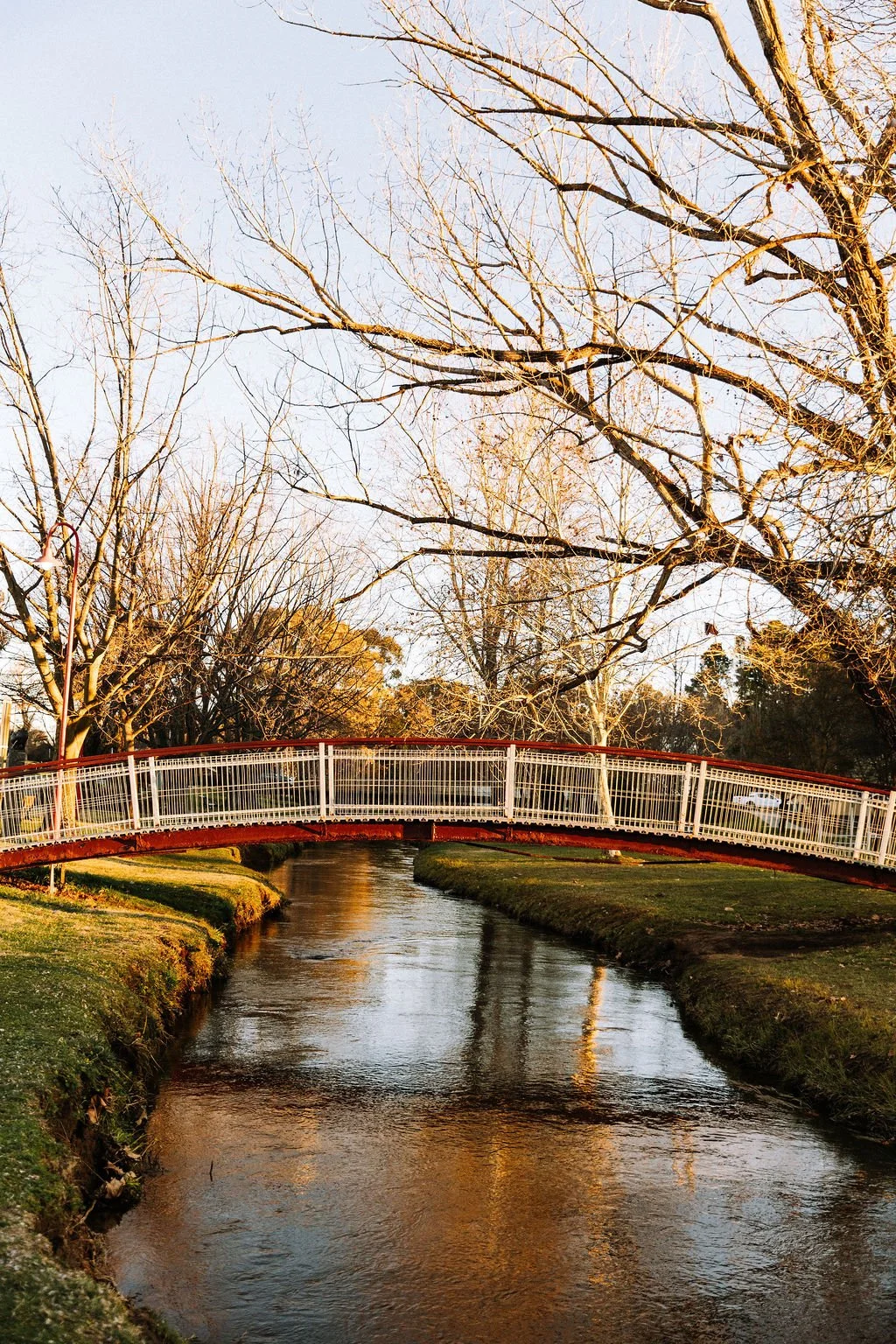 A small, arched, red and white pedestrian bridge crossing over a narrow, calm stream in a park during late fall or early winter, with leafless trees and a partly cloudy sky.
