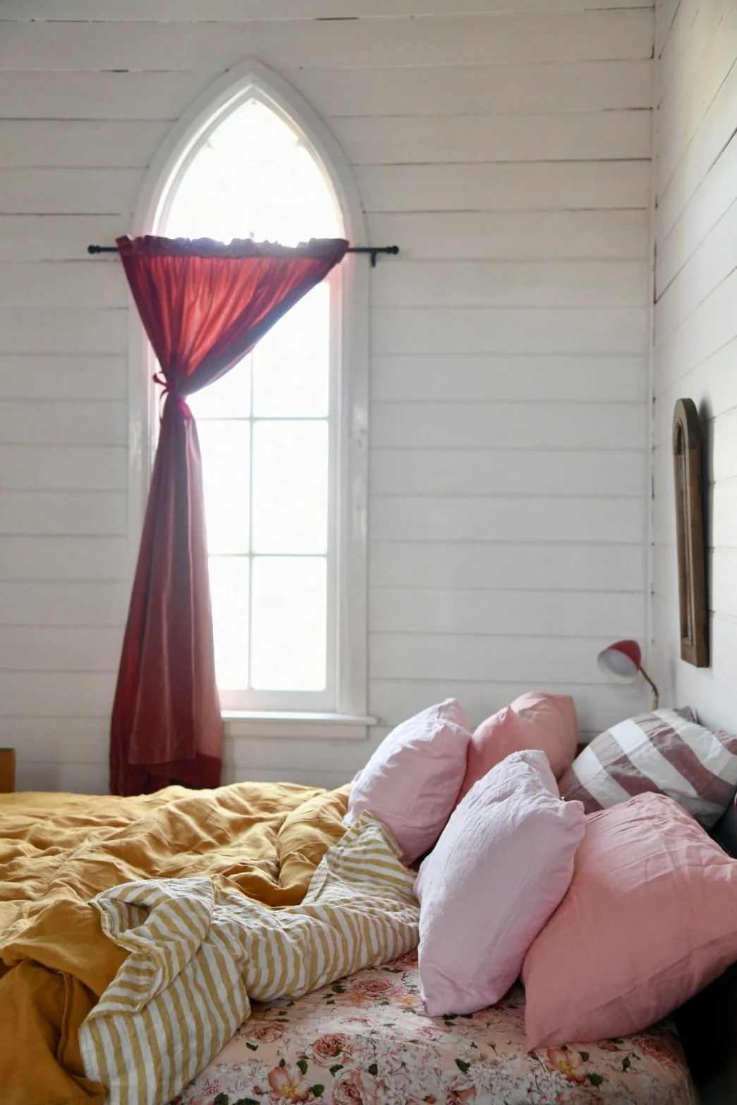 A cozy bedroom with a wooden paneled wall, arched window, red curtains, bed with pink pillows, yellow blanket, and striped linens.