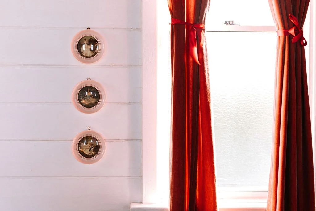 Red curtains tied back with bows hang beside a window with frosted glass, illuminating the room with bright light. Three small round mirrors with pink frames are mounted vertically on a white wall to the left of the window.