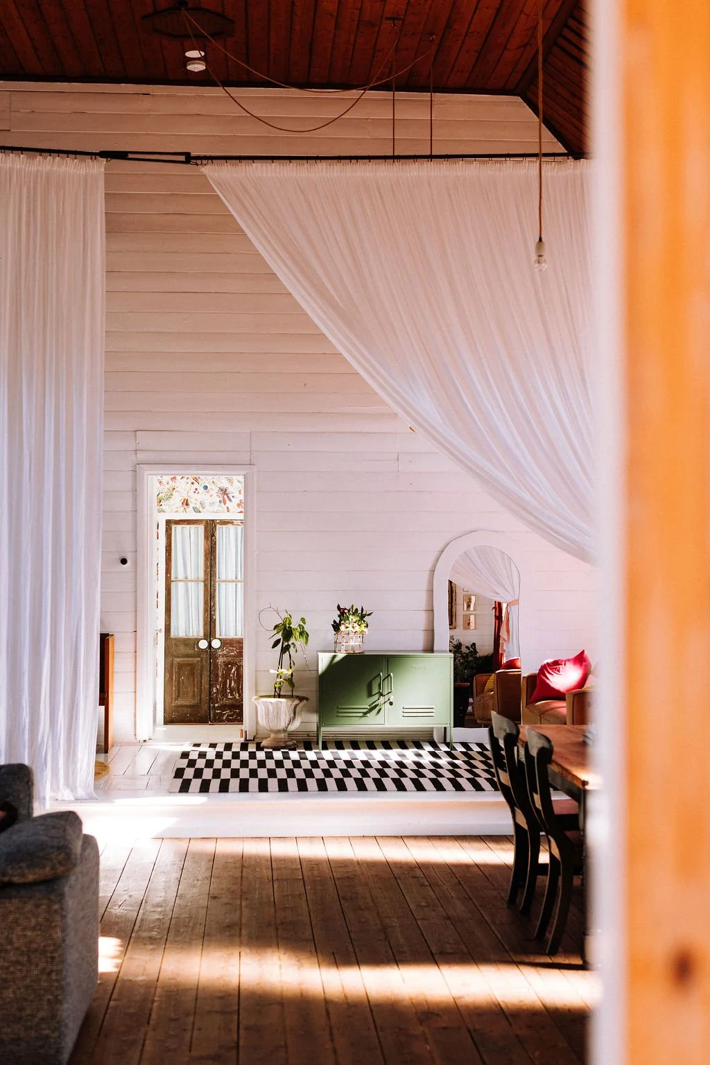 A cozy living room with wooden floors and white shiplap walls, featuring white curtains draped from the ceiling, a large mirror, a green sideboard, red pillows on a sofa, and a checkered black and white rug.