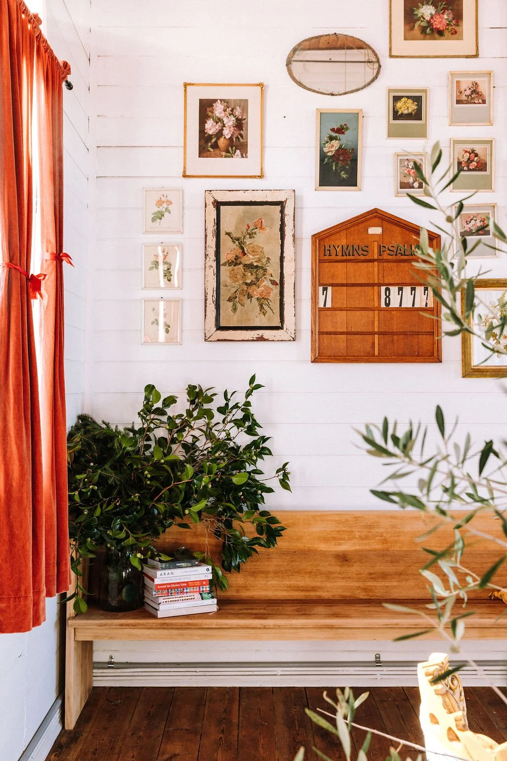 Decorative wall with framed artwork, a wooden hymn board, plant on a wooden bench, and red curtains in a cozy interior.
