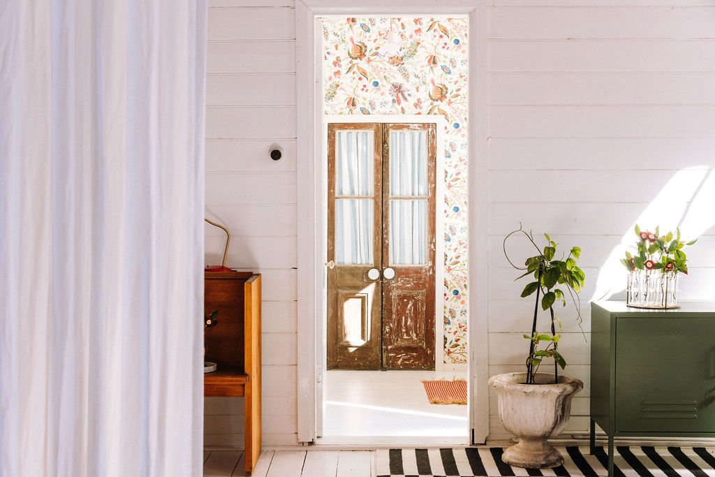Interior view of a room with a white wooden wall, a partially open door with a floral wallpaper border behind it, and two sets of double doors with glass panes. There is a potted plant in a decorative planter, a vintage green cabinet with plants and 
