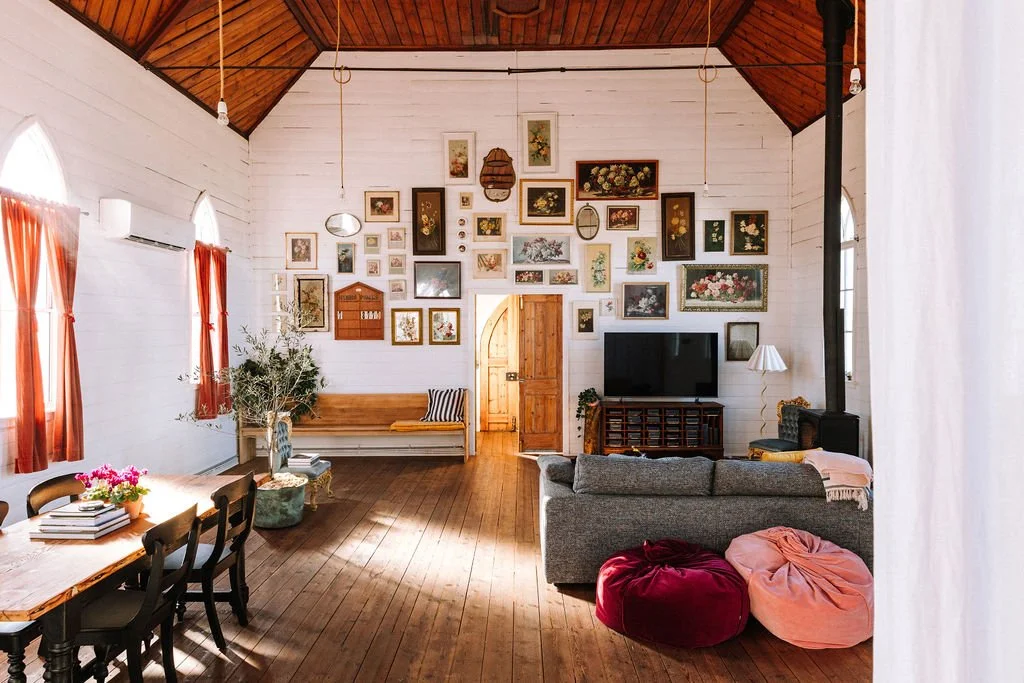 Living room with a wooden high ceiling, white painted wooden walls, and hardwood flooring. Decorated with numerous framed pictures and artwork on the wall. Contains a gray sofa, pink and red bean bags, a wooden bench with cushions, a TV on a stand, a