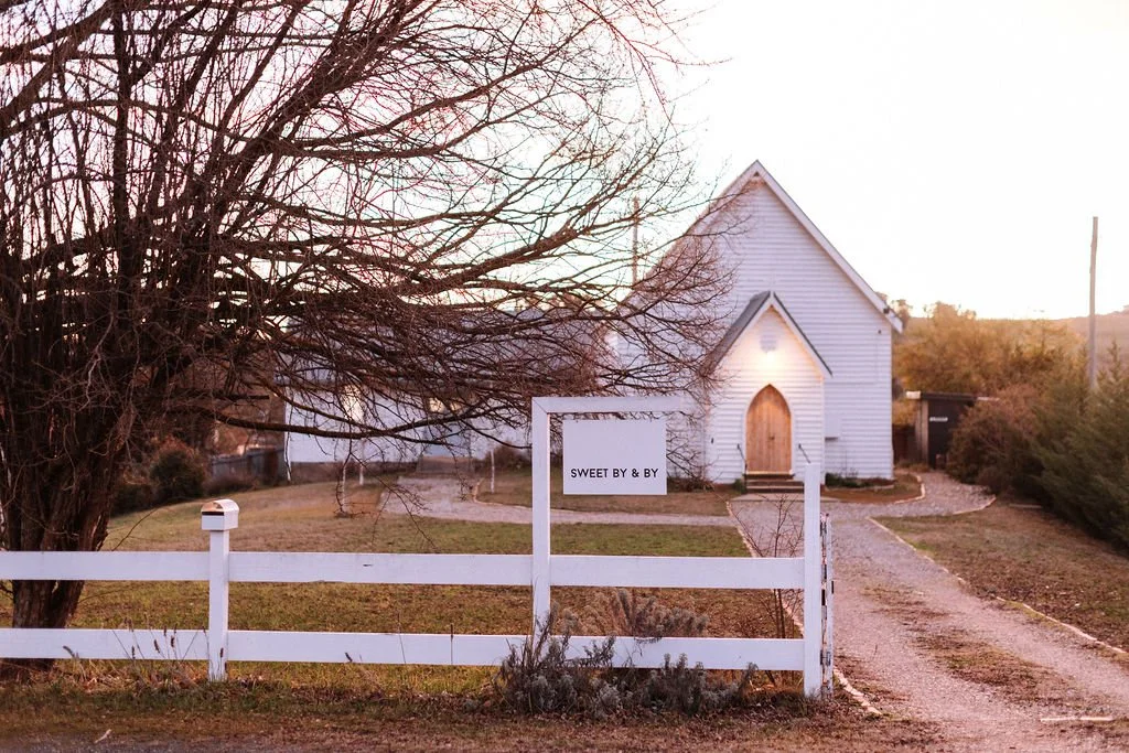 White church with a wooden front door, surrounded by a white fence and a leafless tree, during sunset with soft pink and orange sky.