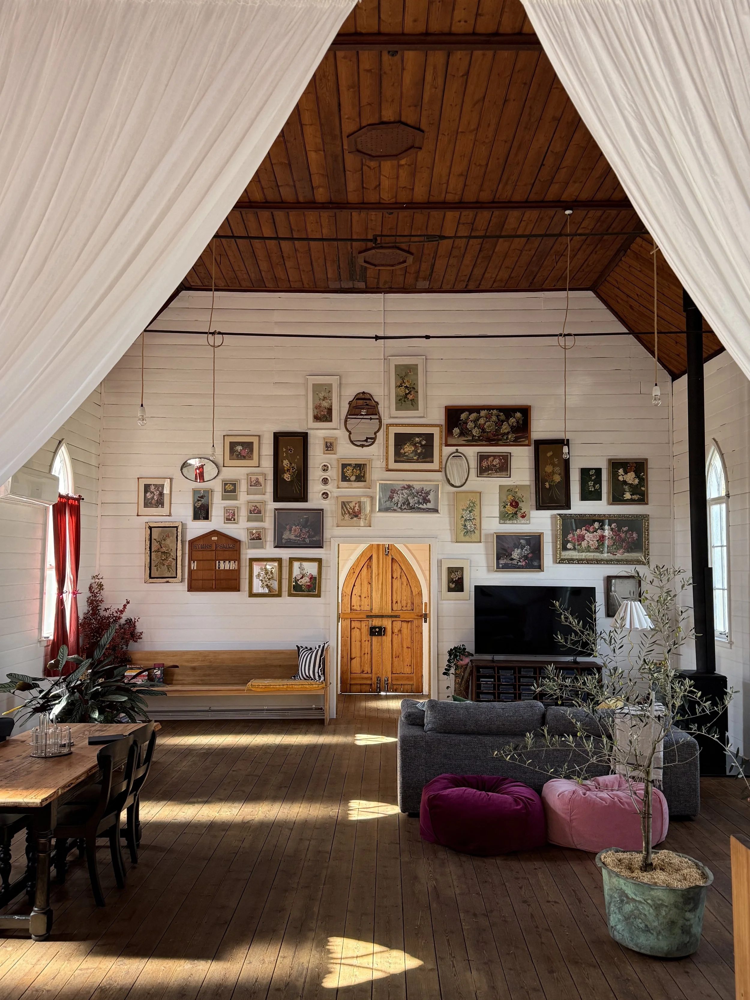 Interior of a converted church with wooden ceiling, white walls, and wooden floor. A mix of modern and vintage furnishings, including a couch, dining table, and poufs. Various framed artwork and photos arranged on the walls. Large arched door at the