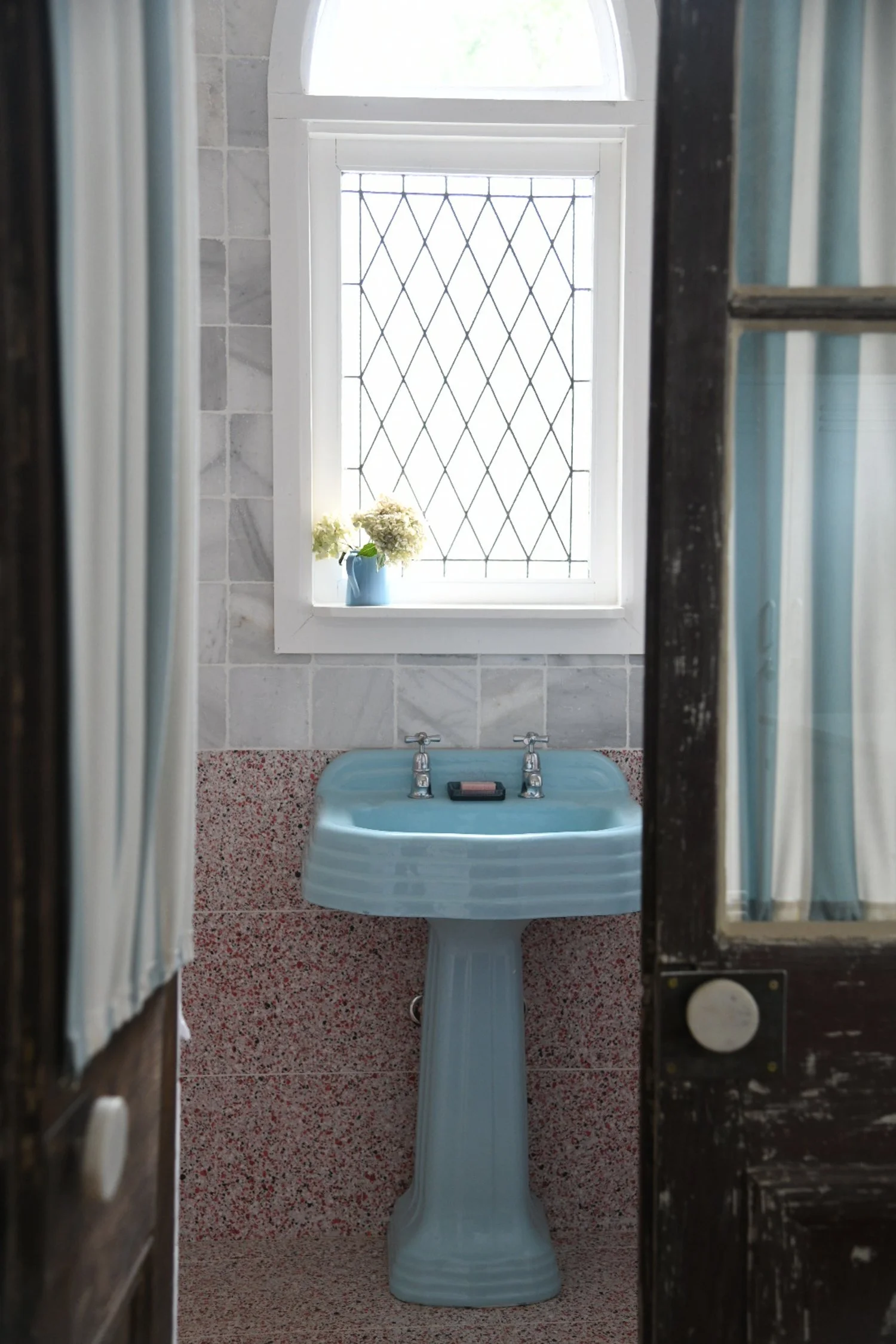Vintage bathroom interior with blue porcelain sink, diamond-patterned window, and flowers in a small vase.