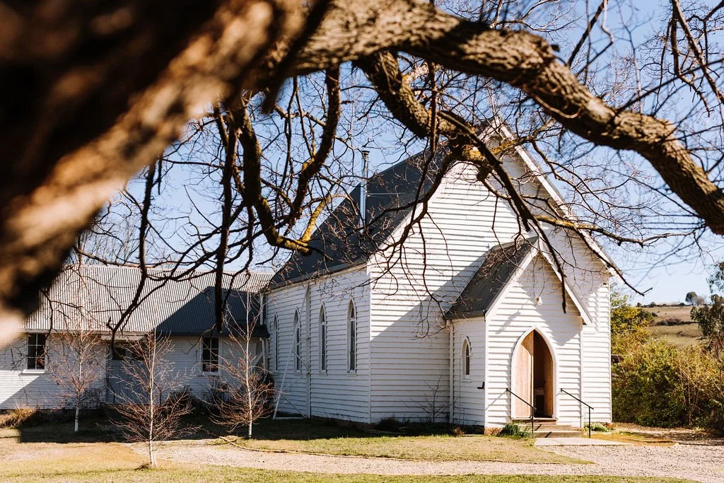 A white wooden church with a steep roof and arched windows, seen through tree branches, in a rural landscape.