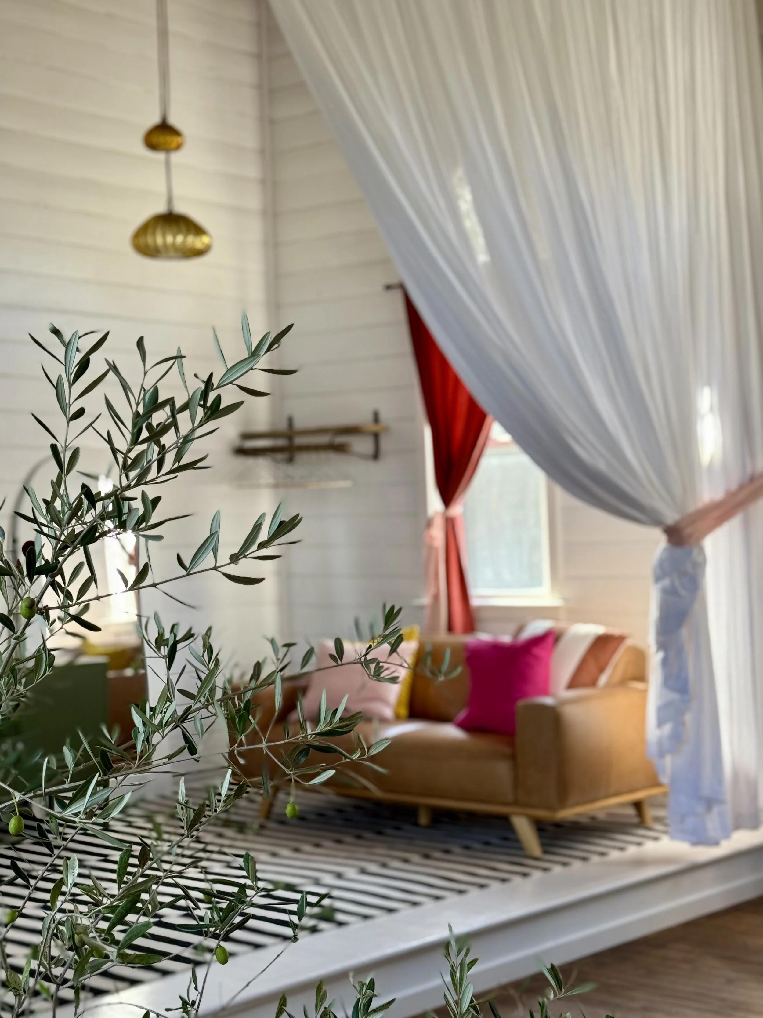 Cozy living room with a brown leather couch, colorful pillows, striped rug, and sheer white curtains. Olive branches in the foreground and pendant lights hanging from the ceiling.