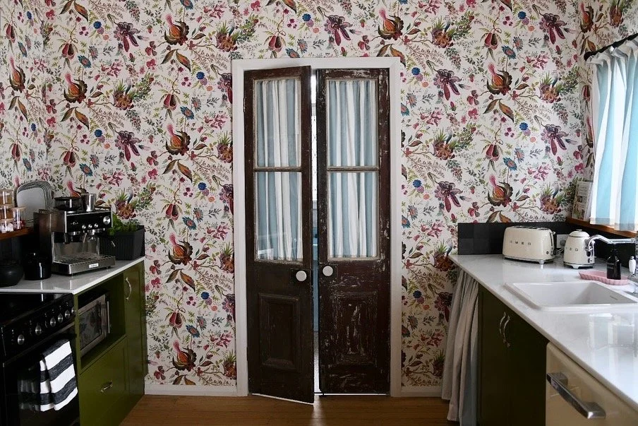 Vintage kitchen with floral wallpaper, wooden doors, coffee maker, green cabinets, white countertops, toaster, and kettle.