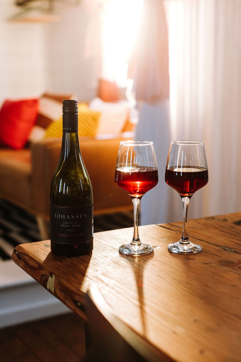 A bottle of pink wine and two glasses filled with red wine are on a wooden table in a cozy living room illuminated by warm sunlight.