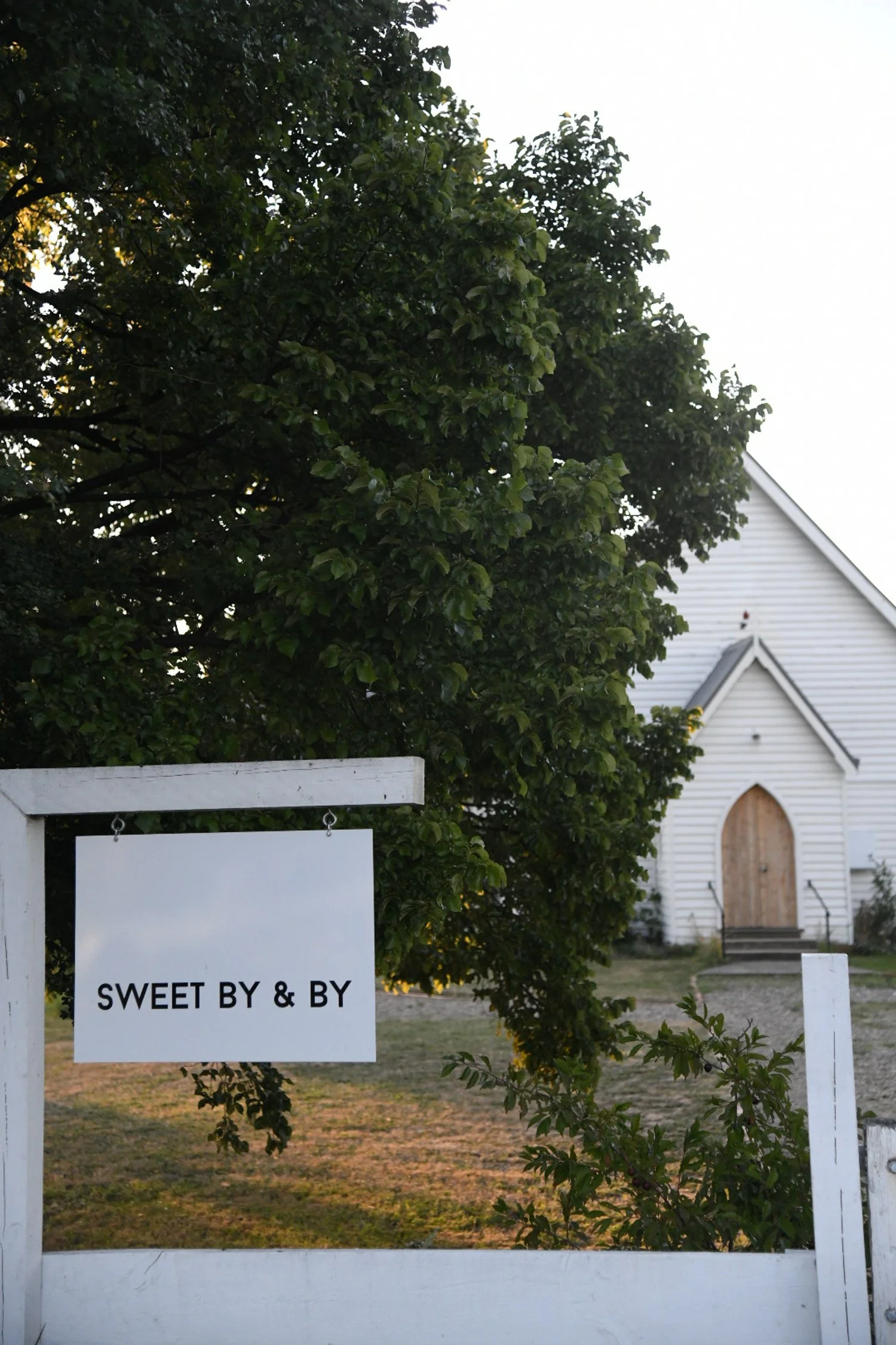White sign reading 'Sweet By & By' hanging on a wooden frame with a white building in the background, partially obscured by a tree.