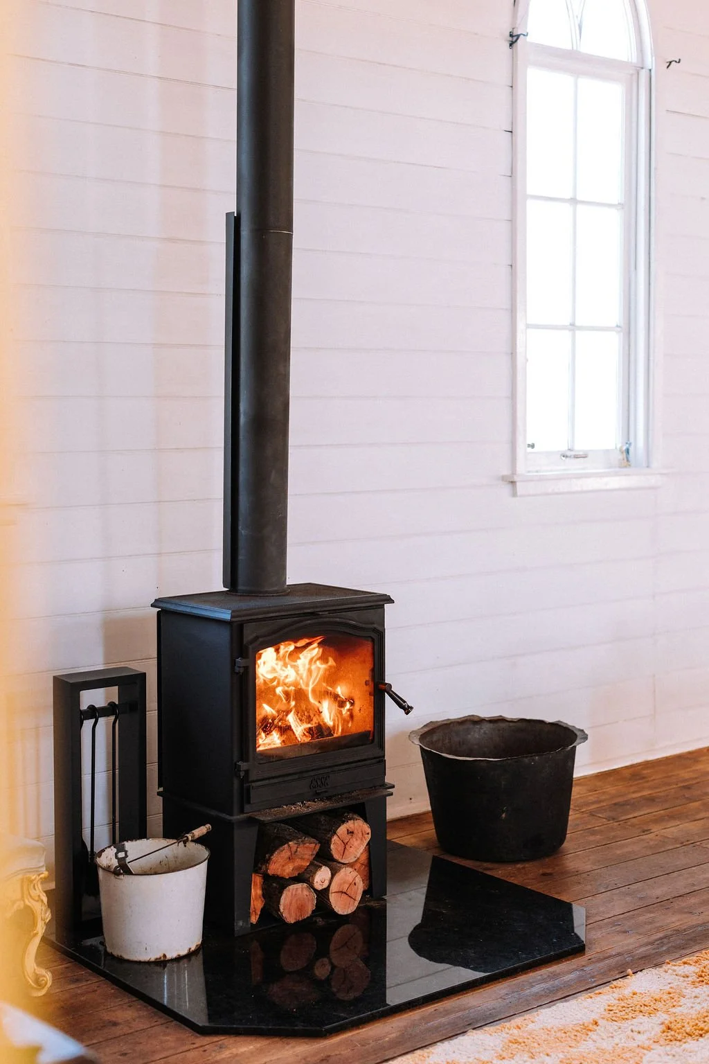 A cozy interior featuring a black wood-burning stove with a fire inside, connected to a black chimney pipe. The stove sits on a black hearth with logs stored underneath. To the right of the stove, there is a large black container, and on the left, a 