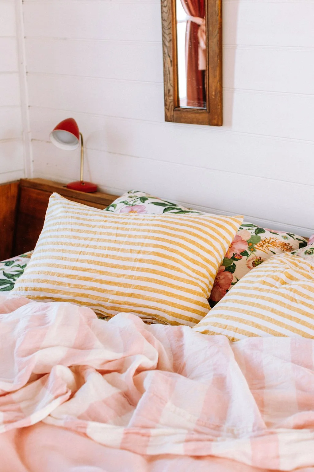 A cozy bedroom scene with a bed featuring striped yellow and white pillows, floral sheets, and a pink checkered blanket. A wooden wall with a mirror and a red bedside lamp are visible in the background.