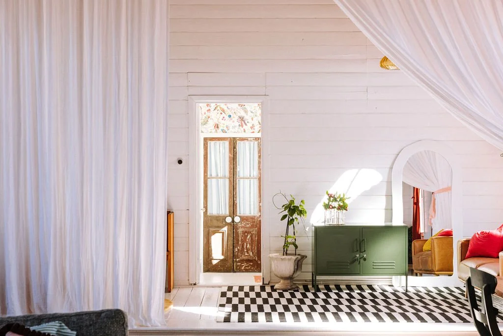 Bright, airy interior of a room with white walls and a black-and-white checkered rug, a green cabinet, a potted plant, a mirror, and a door with frosted glass panels, illuminated by natural sunlight.