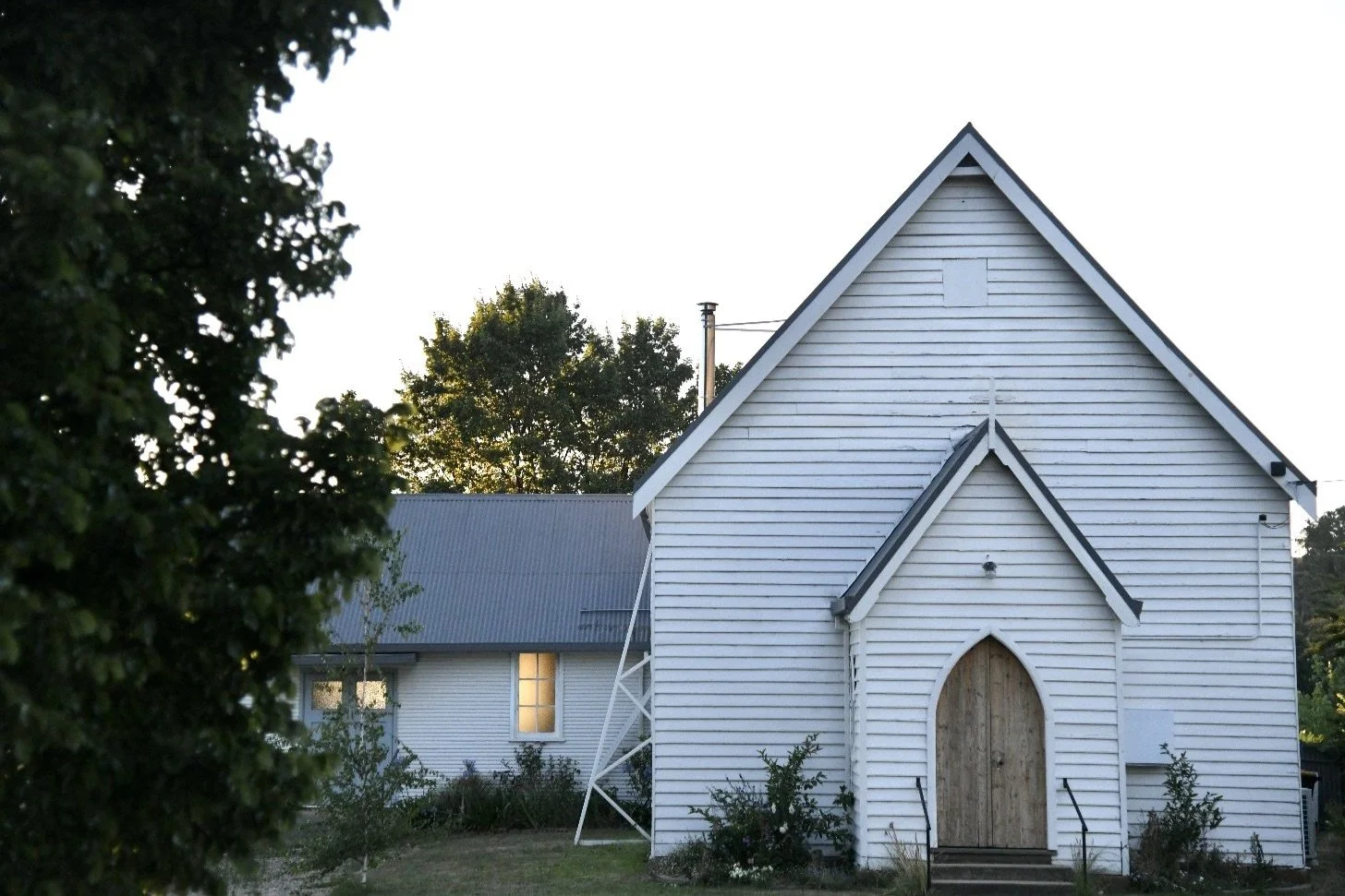White wooden building resembling a church with arched doorway, surrounded by trees and greenery at sunset.