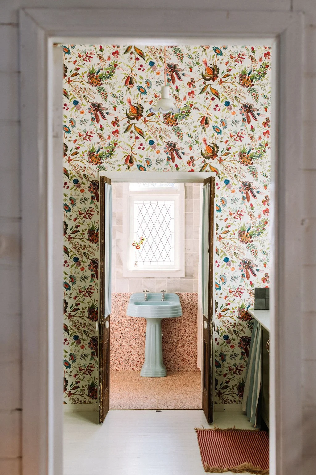 Vintage bathroom with floral wallpaper, a light blue pedestal sink, a window with diamond-shaped leaded glass, and a small rug on white wooden floor.