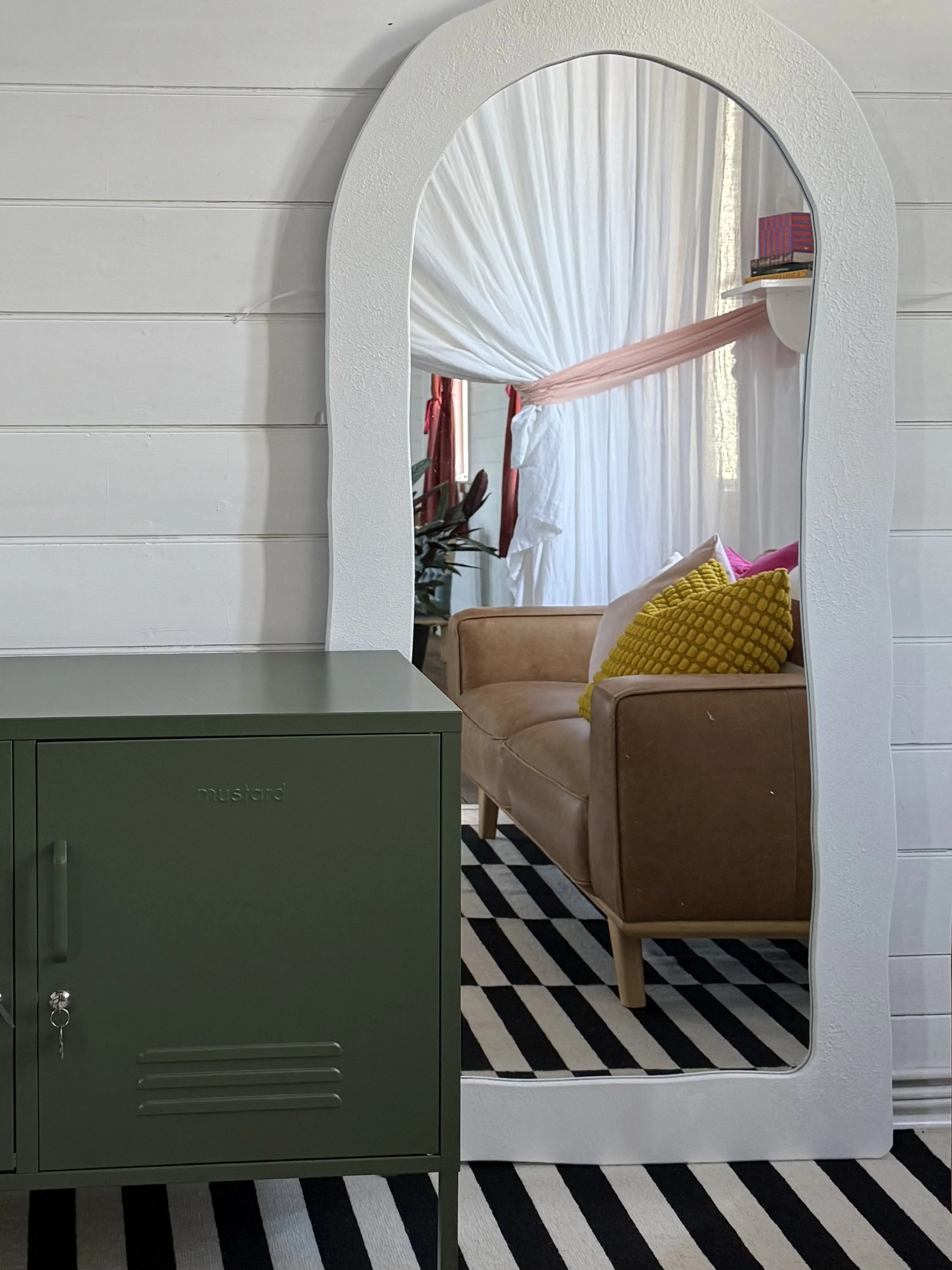 A large white-framed mirror leaning against a white shiplap wall reflects a living room scene with a brown sofa, colorful pillows, and white curtains. In front of the mirror is a green metal cabinet on a black and white striped rug.