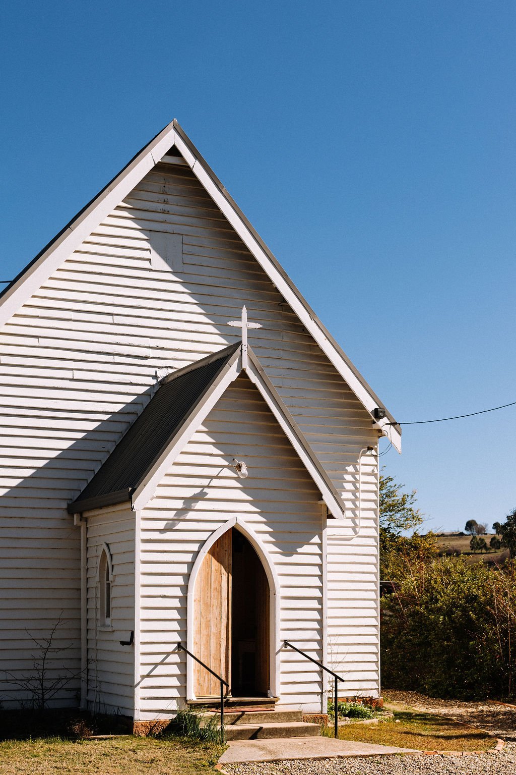White wooden church with arched entrance and pointed roof, set against a clear blue sky and surrounded by trees and bushes.