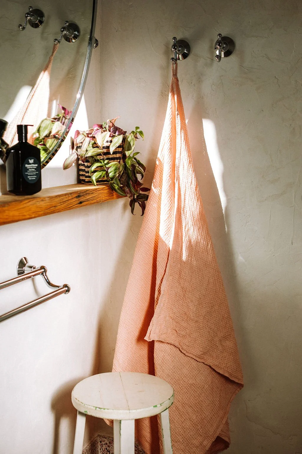 A bathroom corner with a peach-colored towel hanging on a wall hook next to a small white stool, a shelf with a plant in a striped basket, a soap bottle, and a mirror reflecting part of the towel and hooks.