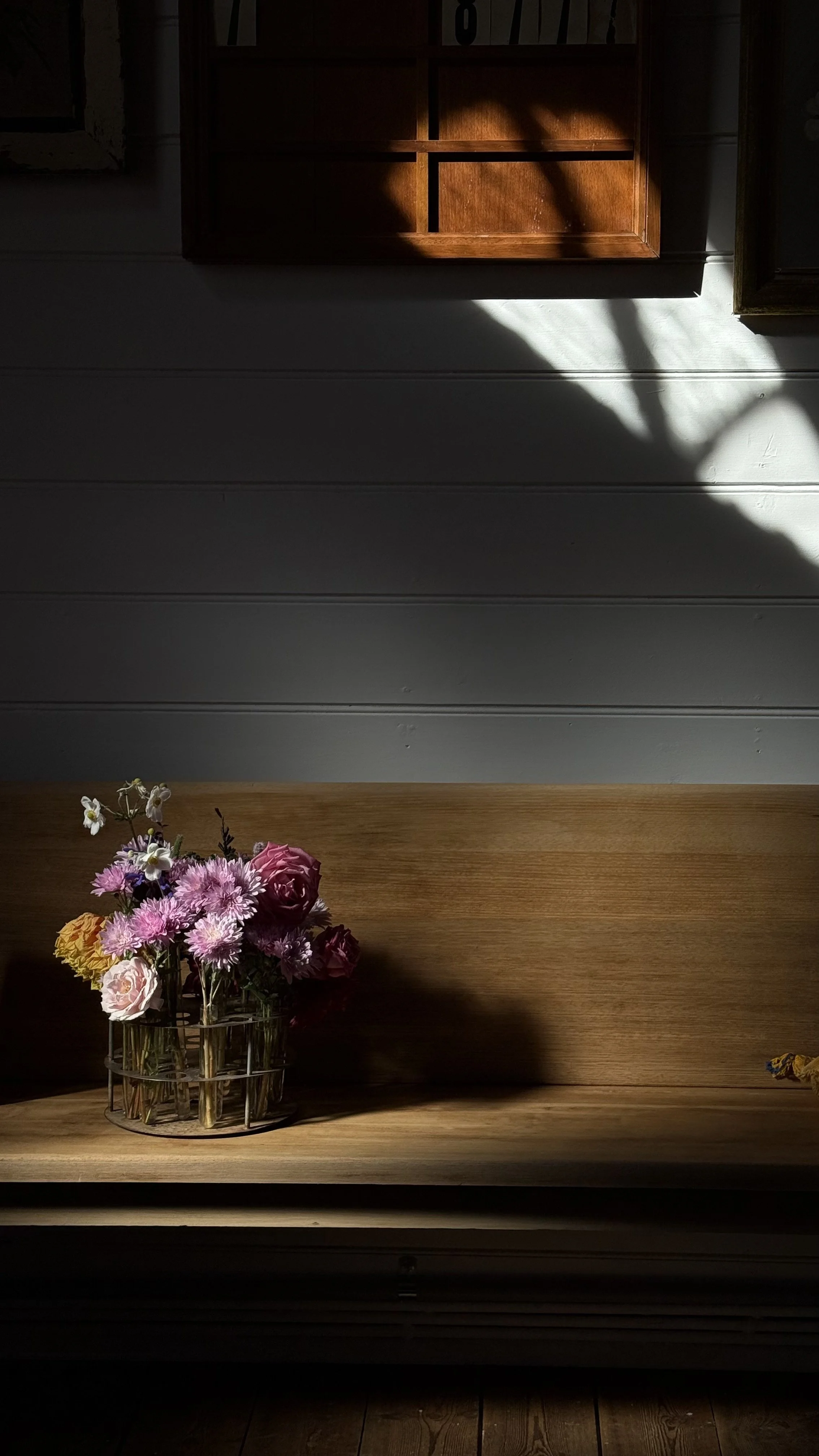 Vase with purple and pink flowers on a wooden table, with sunlight casting shadows on a white paneled wall.