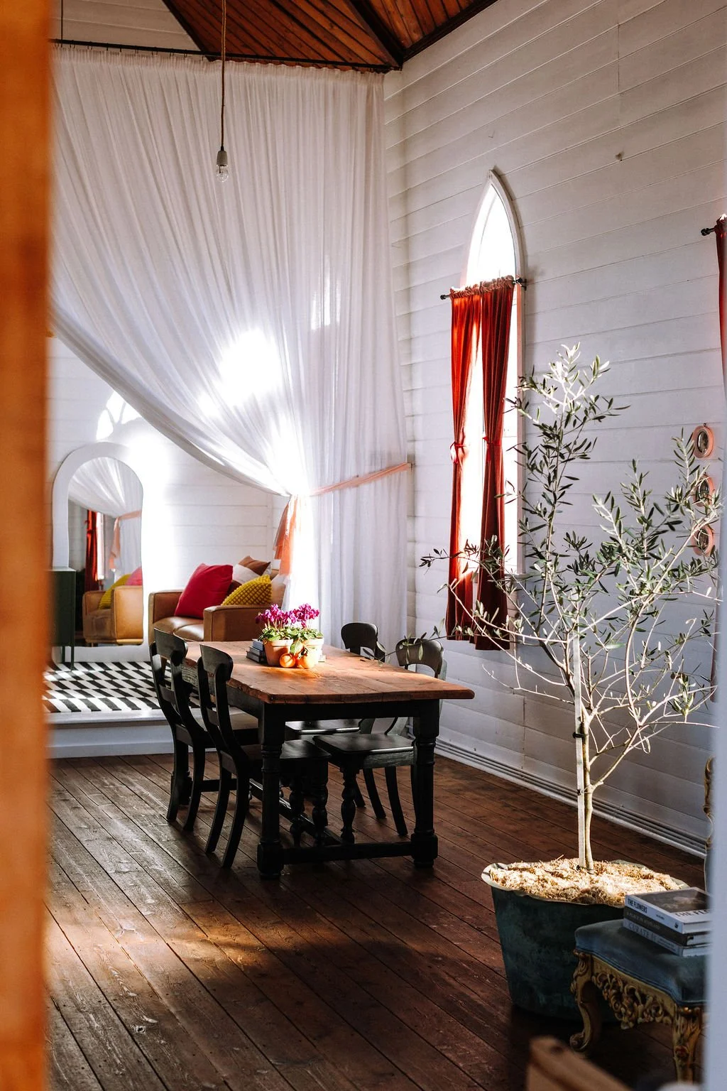 A cozy dining and living area with natural light, a wooden dining table with six black chairs, a large potted olive tree, and a white sofa set with colorful cushions, white drapes, and red curtains.