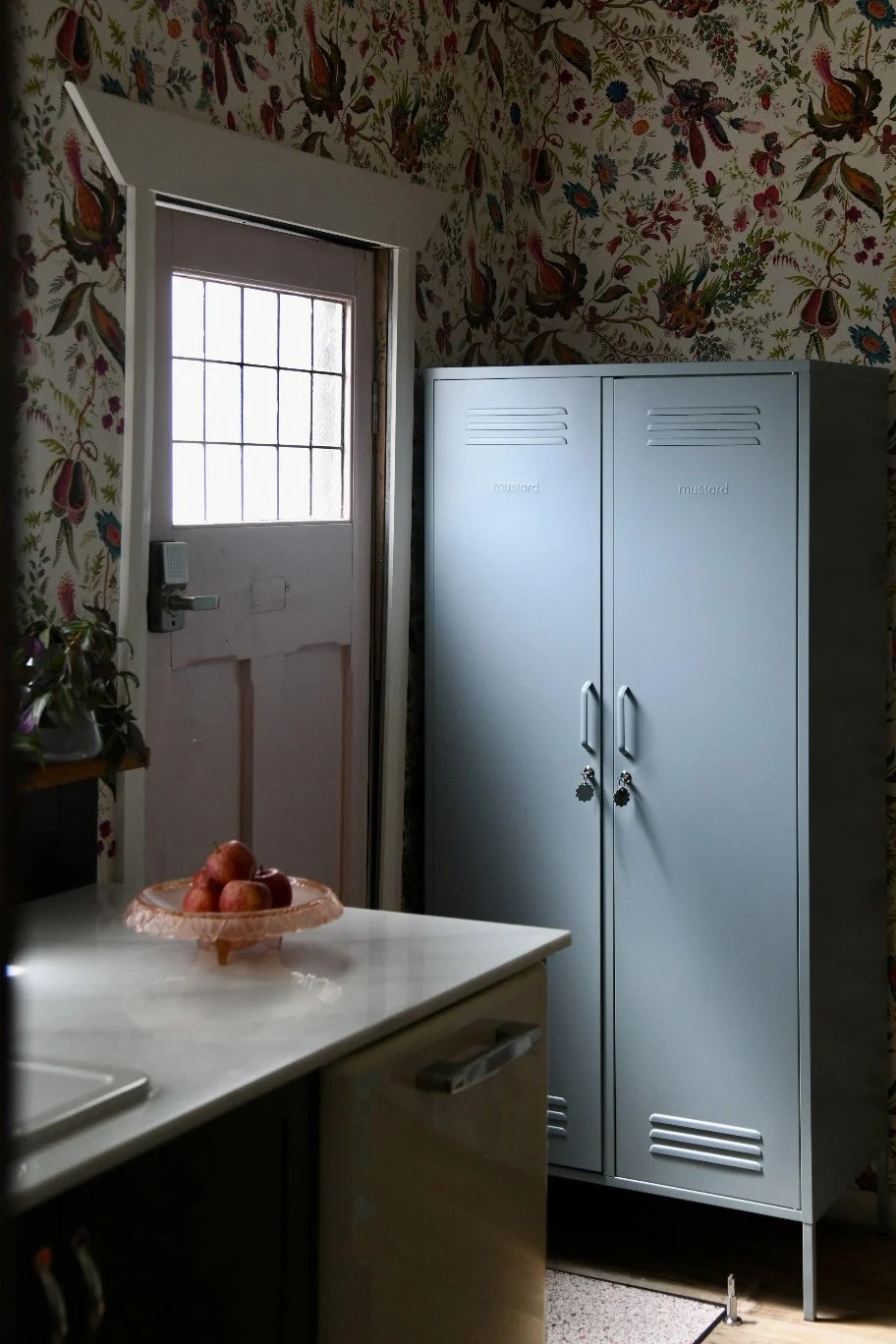 Interior with floral wallpaper, a light blue locker, a fruit bowl with apples on a countertop, and a door with a small window.