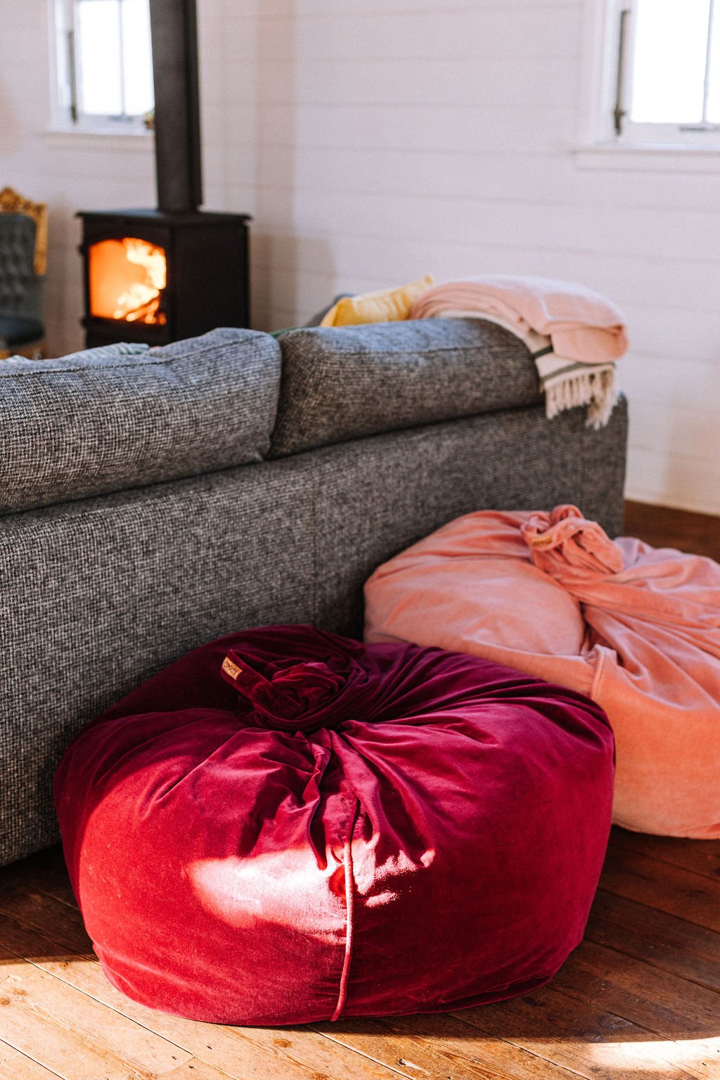 Large red and pink velvet bean bag chairs on a wooden floor in front of a gray sofa in a cozy living room. A stove with visible fire is in the background near a white wall with two windows.