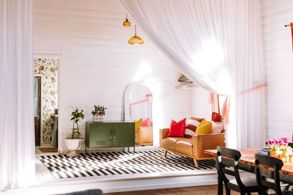 Bright living room with white wooden walls, a black and white striped rug, a small brown leather sofa with multicolored pillows, a green cabinet, a mirror, hanging woven lamps, and a dining table with chairs.