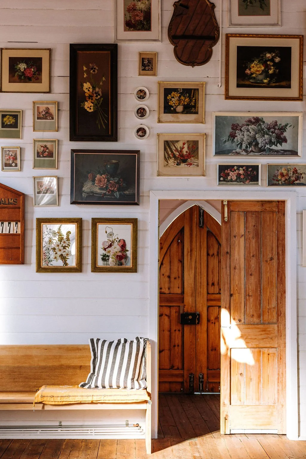 A wall decorated with various framed floral art and small round decorative plates, a wooden arched doorway, and a wooden bench with a striped pillow.